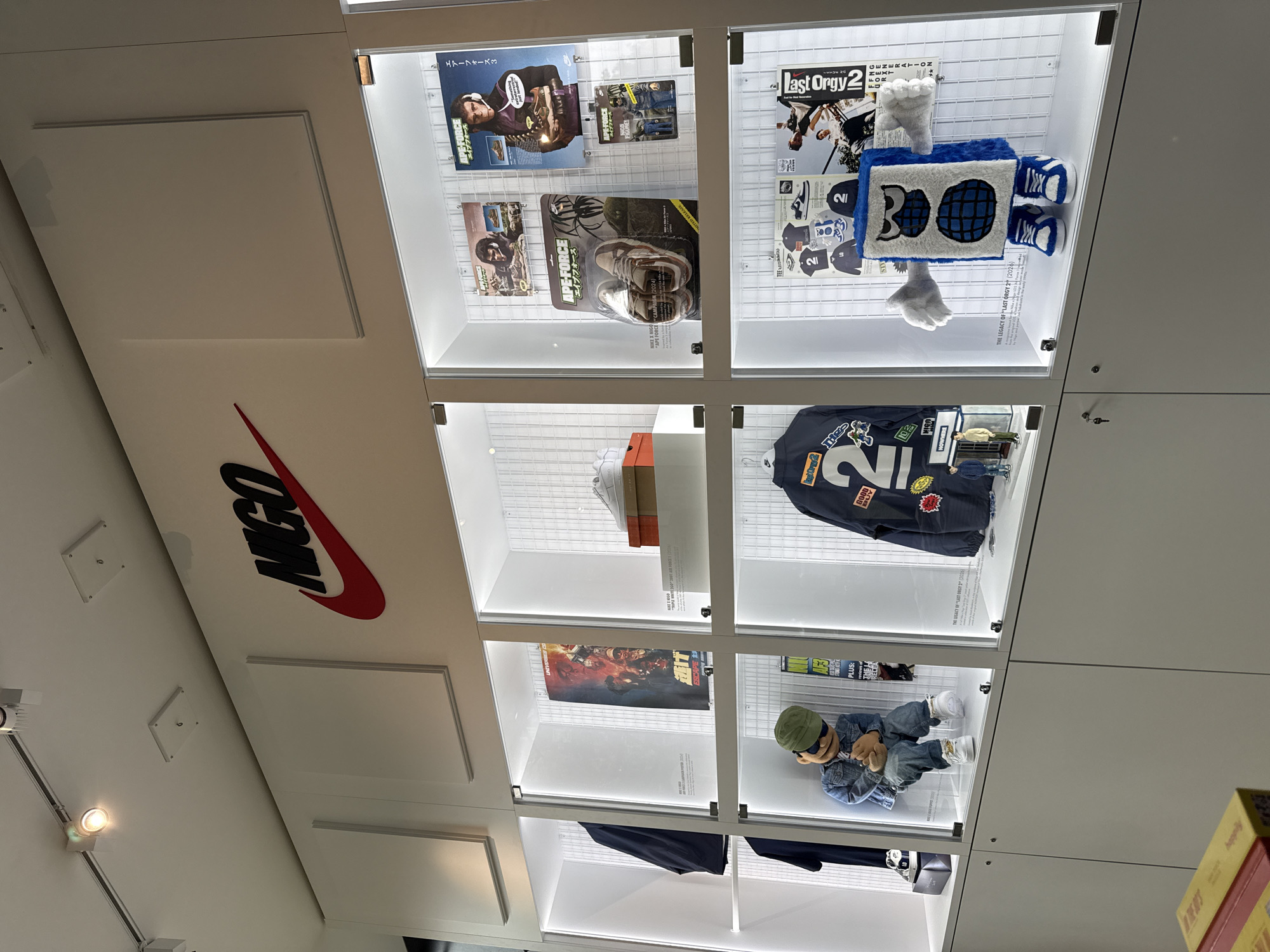 This image depicts a bright, contemporary indoor display space, likely a retail store or exhibition in London, United Kingdom. The scene is dominated by a grid of well-lit glass display cases, showcasing various items related to fashion, streetwear, and collaborations.
Prominently featured on a white wall to the left is a large red Nike swoosh, adjacent to the bold black letters "NIGO," indicating a collaboration with the Japanese designer Nigo.
The display cases contain a variety of artifacts:
The top-left case features merchandise and posters for "APE-FORCE" (エイブフォース), with text mentioning "NIKE X NIGO 'APE FORCE CAMPAIGN (2024)", "ESCA", and "エアーフォース3" (Air Force 3).
The top-right case displays a blue, block-shaped plush toy with sneakers and the number "2", alongside posters for "Last Orgy 2" and text about "THE LEGACY OF LAST ORGY 2 (2026)", "NEXT GENERATION", "TEE", "GOOD", and "NOWHERE".
The middle-left case holds a white Nike sneaker and a shoebox, with visible text "TRIPLE WHITE 260 3001 AIR FORCE 1".
The middle-right case exhibits a dark denim jacket decorated with patches, including "GOOD BOY" and "NOWHERE", and the number "2".
The bottom-left case contains a poster, possibly for a movie or game, with "PLUS: THE H" visible.
The bottom-right case features a small mannequin wearing a green cap, denim jacket, jeans, and white sneakers, with text referencing "THE LEGACY OF LAST ORGY 2+ (2026)".
The setting is meticulously clean and modern, characterized by white walls, overhead track lighting, and transparent display cases that illuminate the items, presenting them as curated pieces of cultural and fashion history.