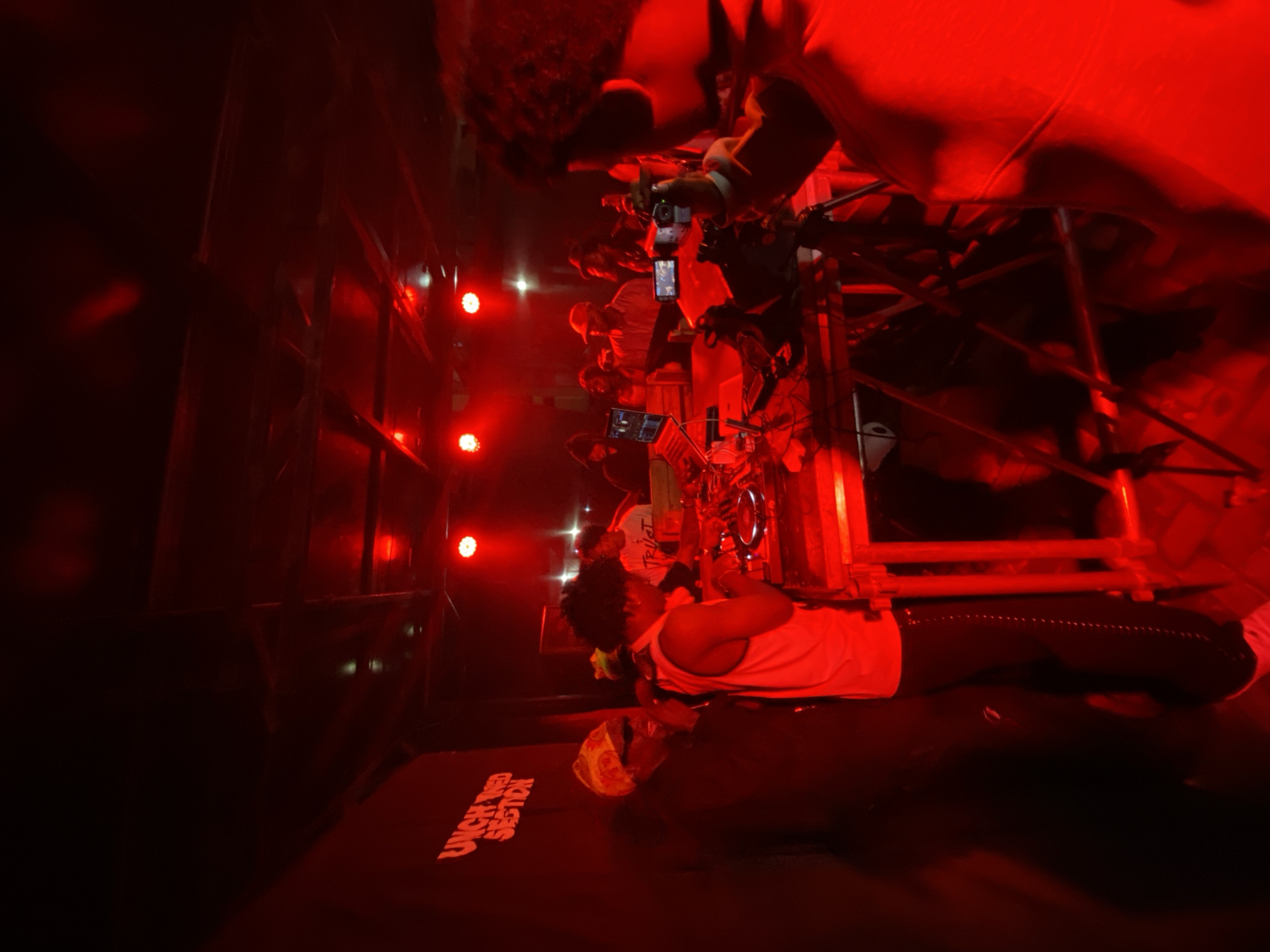 In Abuja, Nigeria, a dimly lit event unfolds under the vibrant glow of red stage lighting. A backdrop on the left displays the words "UNCHAINED SECTION" in bold, orange lettering. In the foreground, a person wearing a white tank top and black pants stands near a DJ setup, appearing to be operating equipment. To their left, another individual is seen wearing sunglasses and a colorful headscarf. Behind them, a crowd of people is visible, some gathered around tables with laptops and cameras, suggesting a performance or presentation. The overall atmosphere is energetic and social, with the red lighting casting a dramatic hue over the scene.