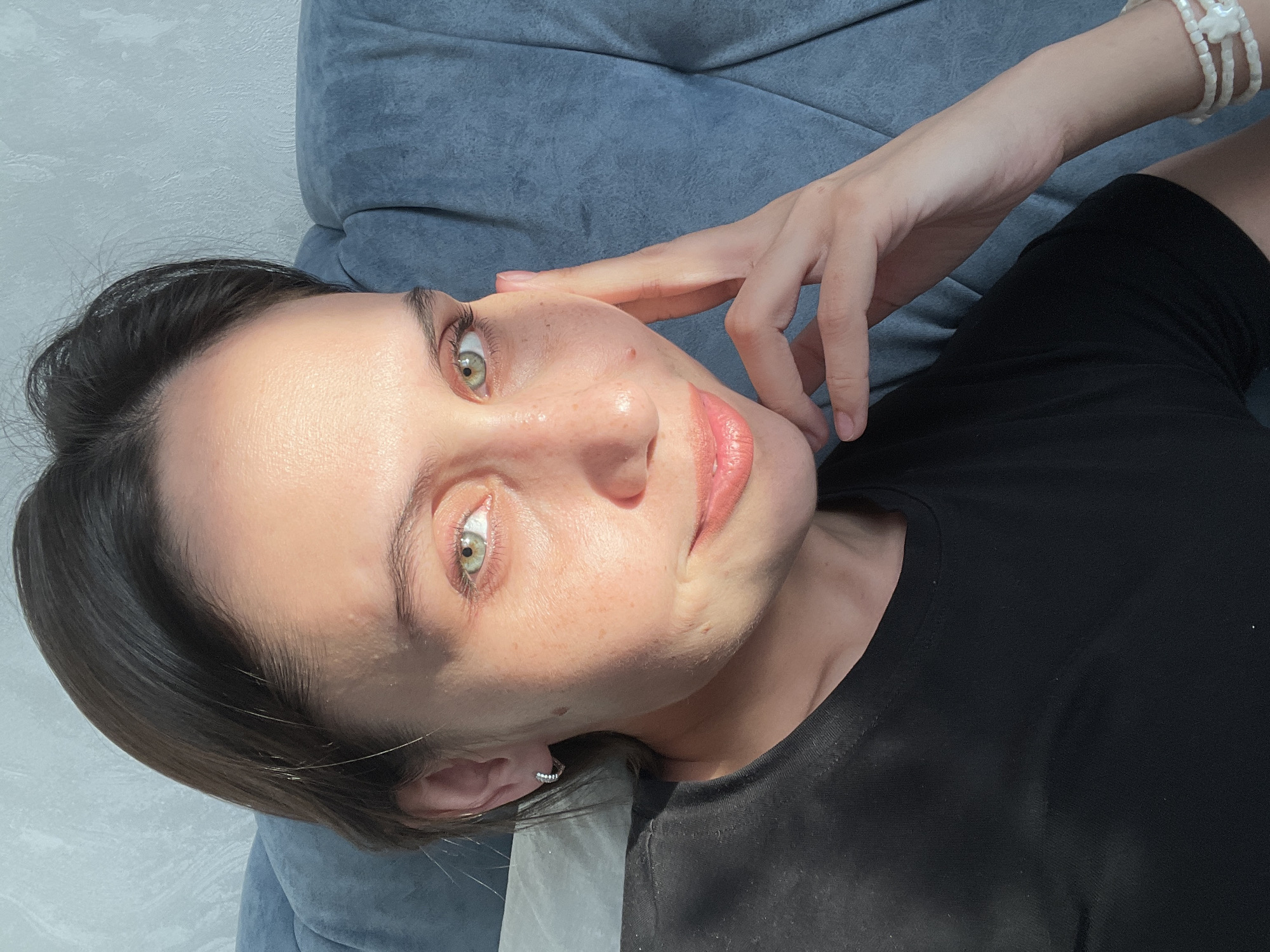 A medium close-up shot depicts a woman with light skin, freckles, and green eyes. She has dark hair styled back from her forehead. Her right hand is raised, with her fingers lightly touching her face. The woman is wearing a black t-shirt and a pearl bracelet is visible on her wrist. She is lying on a blue cushion. The background includes a textured white wall. There is no visible indication that it is in Izhevsk, Russia.