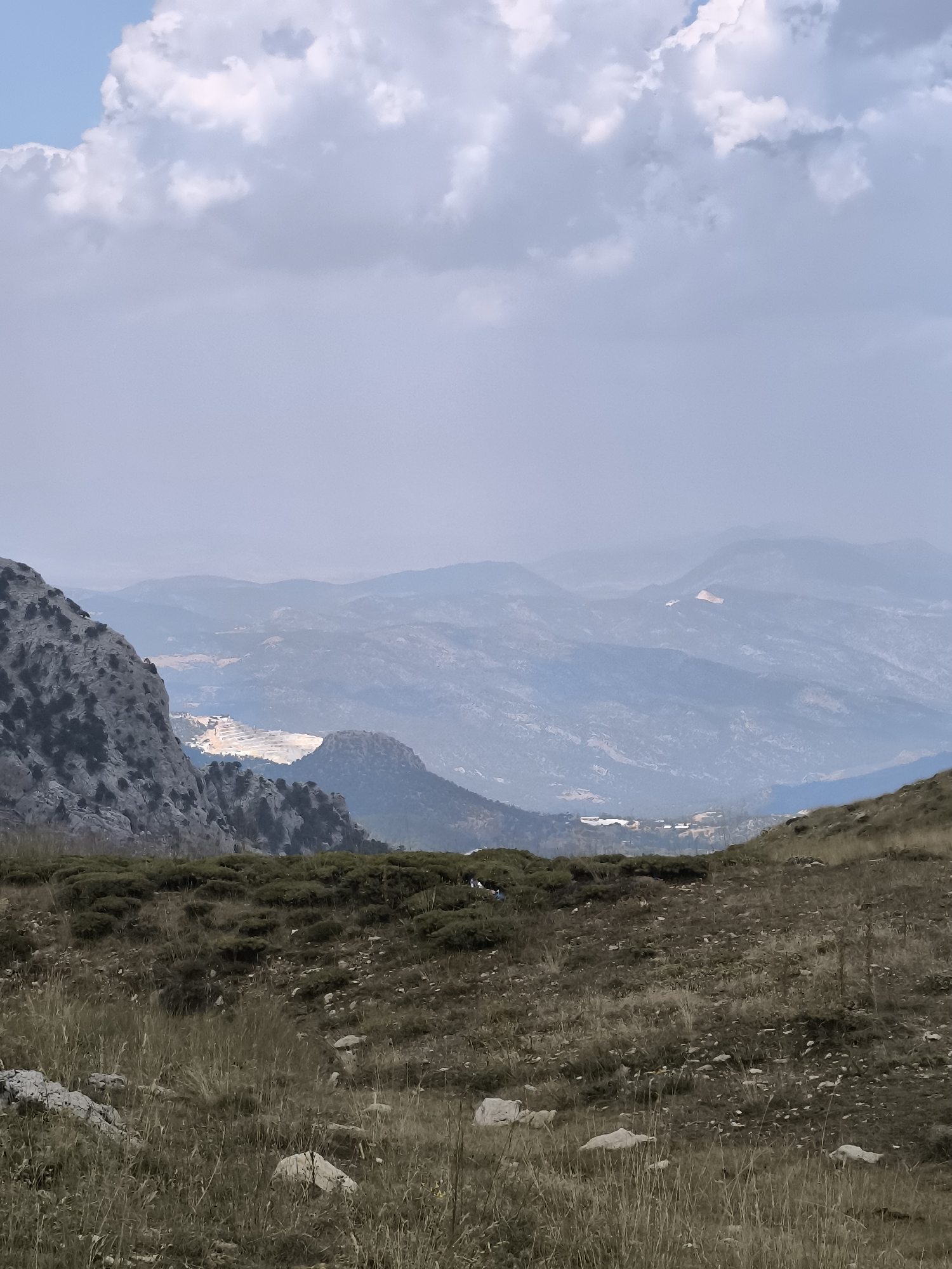 A panoramic view captures the layered, hazy mountain ranges of Karçukuru, Turkey under a partially cloudy sky. In the foreground, a rough, rocky slope covered with dry grass and low-lying shrubs leads the eye towards the distant, undulating landscape. A prominent, grey, rocky outcrop dominates the left side of the frame, its texture rough and weathered. Further in the mid-ground, a large, pale quarry or excavation site is visible, its stark, light-colored earth contrasting with the surrounding muted greens and blues of the mountains. Some structures, possibly buildings, are scattered in the distance. The overall atmosphere is one of vastness and natural beauty, slightly obscured by atmospheric haze.