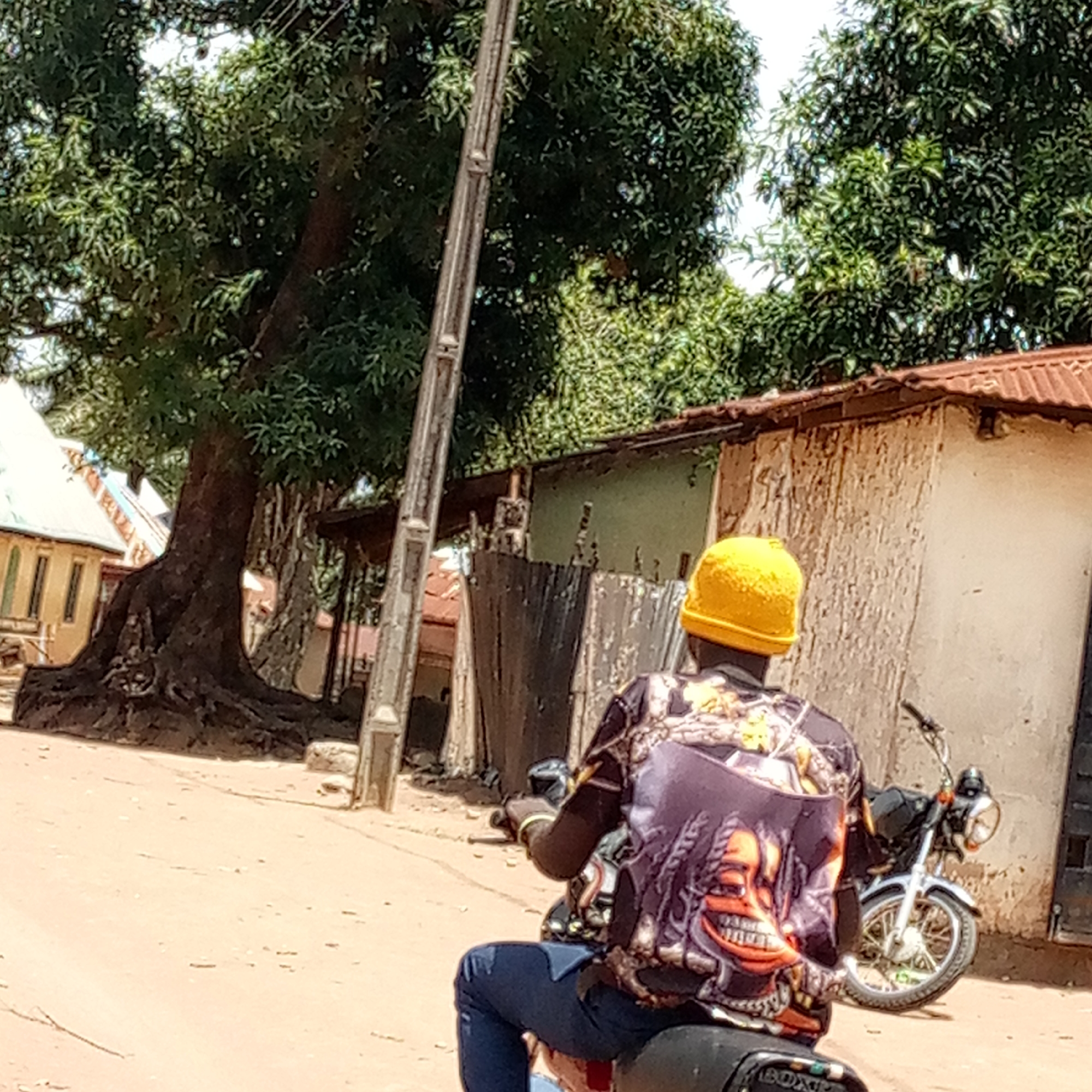 This image captures a daytime scene in Mararaba, Nigeria, showing a person from behind, riding a motorcycle on a light-brown dirt road. The individual wears a bright yellow knitted cap, a long-sleeved shirt with a distinctive purple/black pattern featuring an abstract orange, grinning face, and blue jeans.
To the right of the rider, another motorcycle, dark with silver/chrome accents, is parked near a simple building with a reddish corrugated metal roof and light-colored walls. In the midground, several rudimentary structures and wooden fencing line the dirt path. On the left side of the frame, a very large tree with prominent, exposed roots dominates the landscape, its dense green foliage extending high into the background. A utility pole stands vertically near the tree. Further back, more lush green trees are visible against a bright sky, suggesting a sunny day. The overall impression is that of a bustling, semi-urban or rural area under bright natural light. Partial text "BOXER" or "BOX" is visible on the rear of the rider's motorcycle.