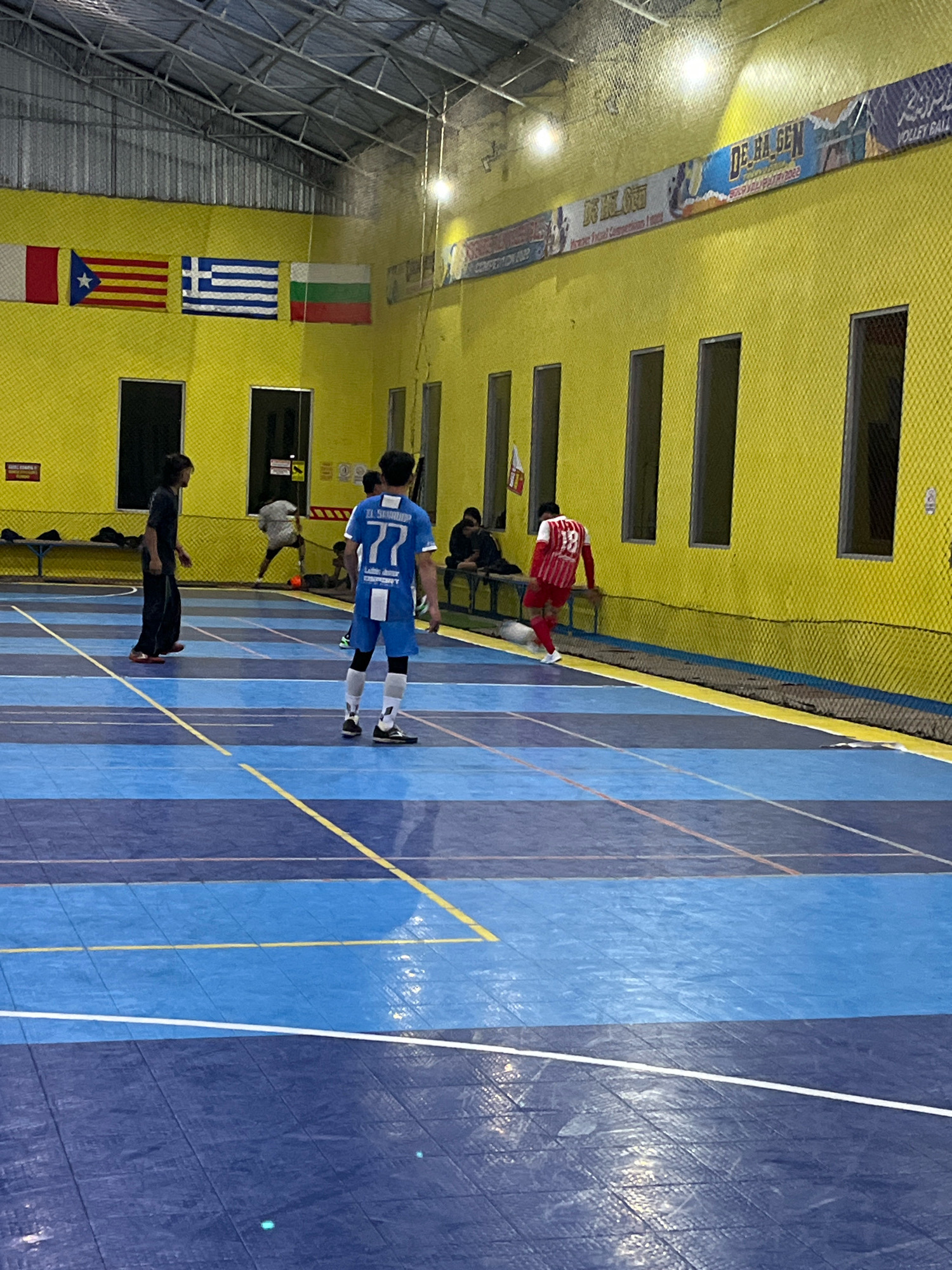 An indoor futsal court in Jatilawang, Indonesia, is depicted, characterized by its blue and dark blue tiled floor marked with yellow and white lines. The walls are painted bright yellow, and a metal truss ceiling supports lighting fixtures. Along the upper part of the wall, several flags are displayed, including the flags of Catalonia, Greece, and Bulgaria. A banner with text in Arabic and English, reading "VOLLEY BALL," is also visible.
The scene shows a futsal match in progress. Three players are actively engaged. One player, wearing a blue jersey with the number 77 and "EL SUD" written on the back, stands in the foreground. Another player, in a red and white striped jersey with the number 10, is near the right side of the frame, dribbling a white ball. A third player, in a dark blue shirt and pants, is positioned further back on the left. In the background, other individuals are seated on a bench, and one person is visible tending to something on the ground. The environment appears to be a sports facility, likely a community or recreational center.
