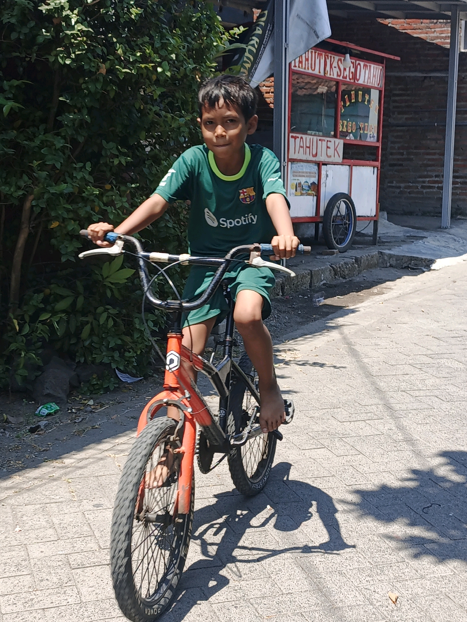 A young boy, appearing to be of Indonesian ethnicity with dark hair and brown eyes, is pictured riding a red and black bicycle on a paved street. He is wearing a green t-shirt with a Spotify logo and green shorts. The boy's left hand is on the handlebars, and his feet are visible, barefoot, resting on the pedals.
In the background, a red and white food cart is visible, with text that appears to read "TAHUTEK-SEGO TAHU" and "TAHUTEK" in Indonesian. A large bush with green leaves is to the left of the boy, and a brick wall is behind the food cart. The ground is paved with interlocking bricks, and shadows from trees and the boy are cast on the surface. The environment suggests a warm, sunny day in a residential or commercial area, possibly within the city of Ketegan, Indonesia, indicated by the signage on the food cart.