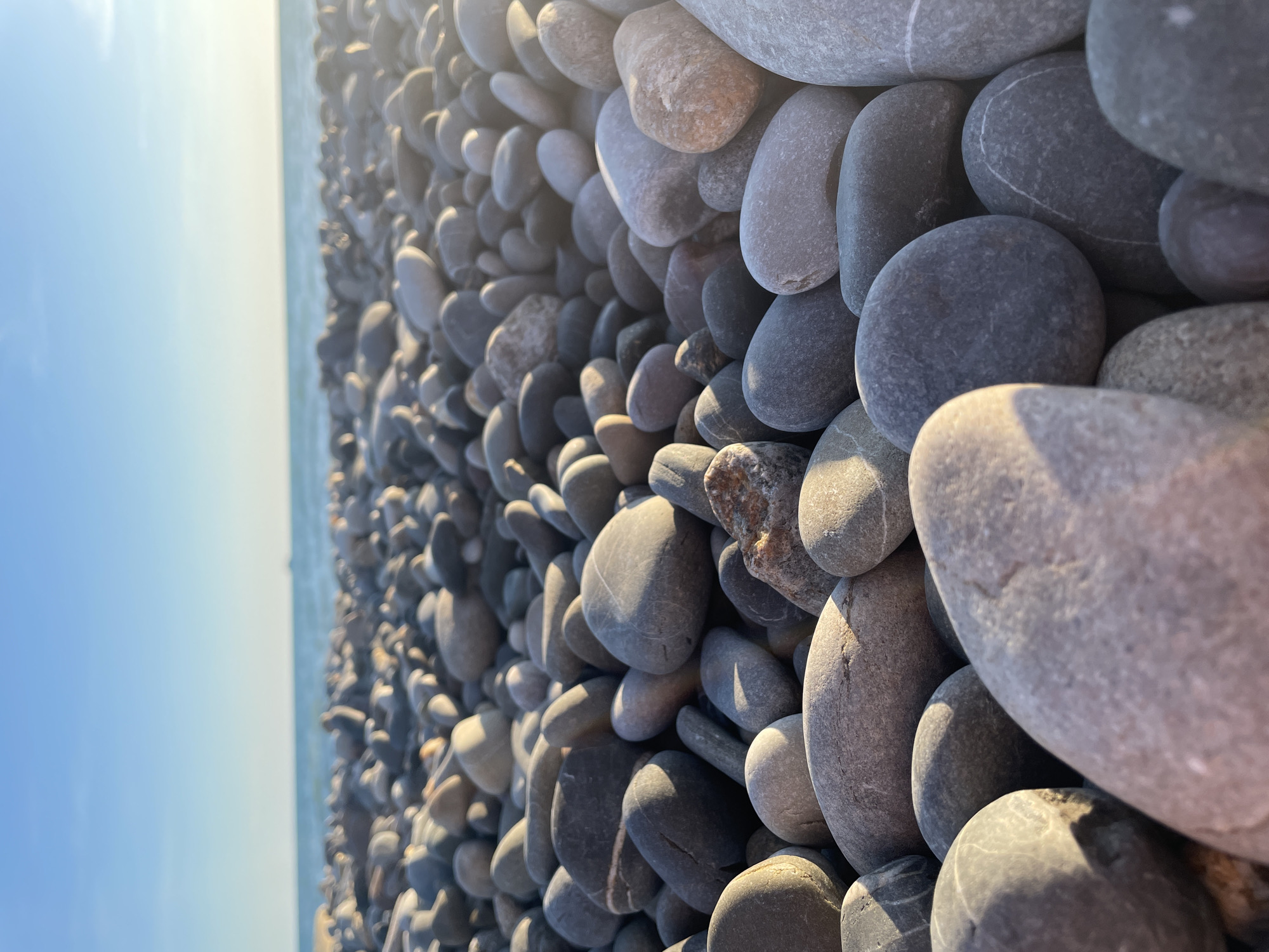 The image depicts a close-up, low-angle view of a pebble beach with the ocean and sky in the background. The foreground is dominated by smooth, rounded pebbles of various shades of gray, brown, and white. Some pebbles have white striations or markings. The pebbles are densely packed and arranged in a way that creates a textured surface.
To the left of the frame, the ocean is visible as a blurred expanse of pale blue or turquoise water meeting a bright, hazy sky. The sunlight suggests it is daytime, likely morning or late afternoon due to the soft lighting and lens flare. The overall mood is tranquil and natural, focusing on the tactile quality of the stones and the serene coastal environment. There is no text visible in the image.