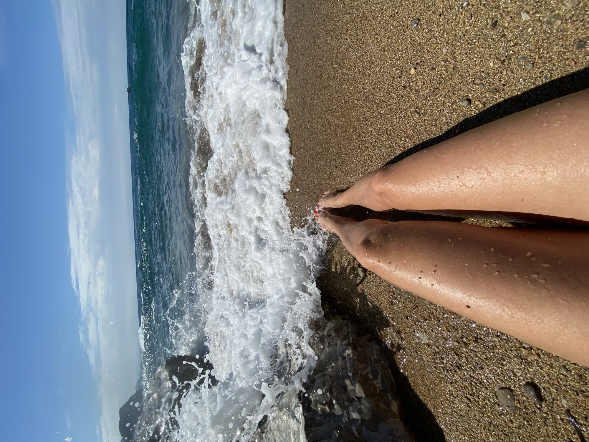 The image, taken at Kamenovo, Montenegro, is a vertical, high-angle shot capturing a relaxing beach scene. The primary subjects are a person's legs and feet, positioned on the right side of the frame, resting on the wet, sandy shore. The legs appear tanned and are dotted with water droplets, while the toes have bright red nail polish and an ankle bracelet is visible on the right foot. A strong shadow from the legs stretches across the sand, indicating direct sunlight.
To the left, the deep blue-green sea extends to the horizon under a clear blue sky with some wispy white clouds. A large, foamy white wave is actively crashing, engulfing dark rocks near the water's edge and creating splashes. In the far distance on the horizon, a small, dark object, possibly a boat or buoy, is faintly visible. The sand is light brown and appears wet and coarse, with small pebbles interspersed. The scene depicts a bright, sunny day at the seaside, evoking a sense of leisure and enjoyment of nature. There is no visible text.