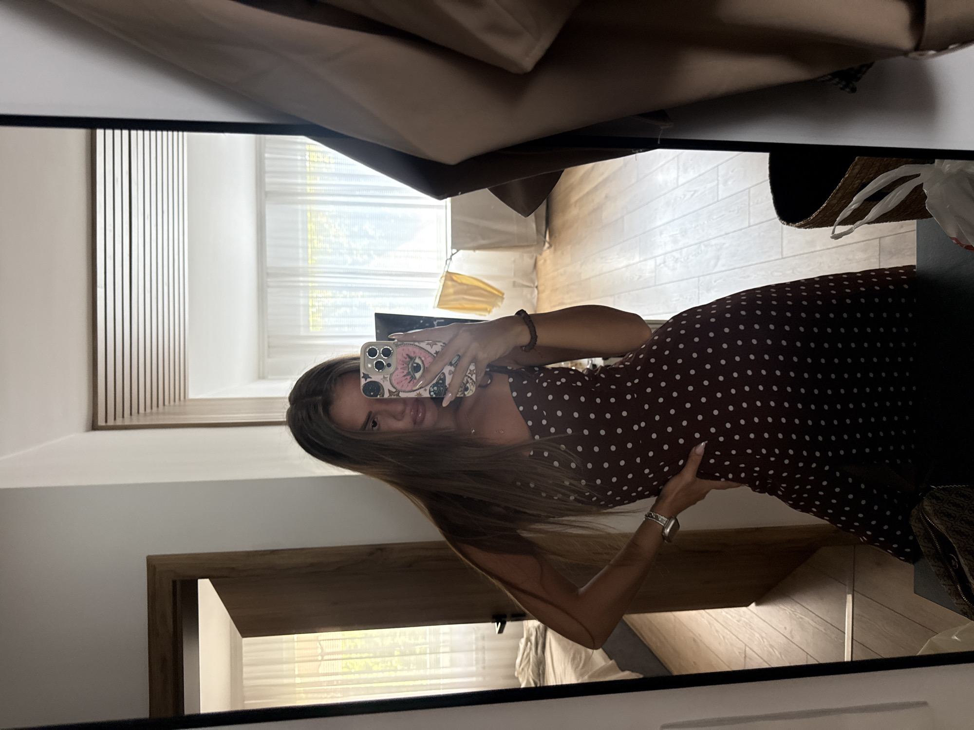 A young woman with long, brown hair is taking a mirror selfie in a bedroom. She is wearing a dark red, polka-dotted dress with spaghetti straps and a silver bracelet on her left wrist. Her phone, with a heart-shaped eye sticker case, is held up to take the picture. The room features light wood floors, white sheer curtains in the background, and a dark wood door. To the right of the woman, a tan coat hangs in the mirror's reflection. A woven basket is visible in the lower right corner. The scene appears to be an interior setting.