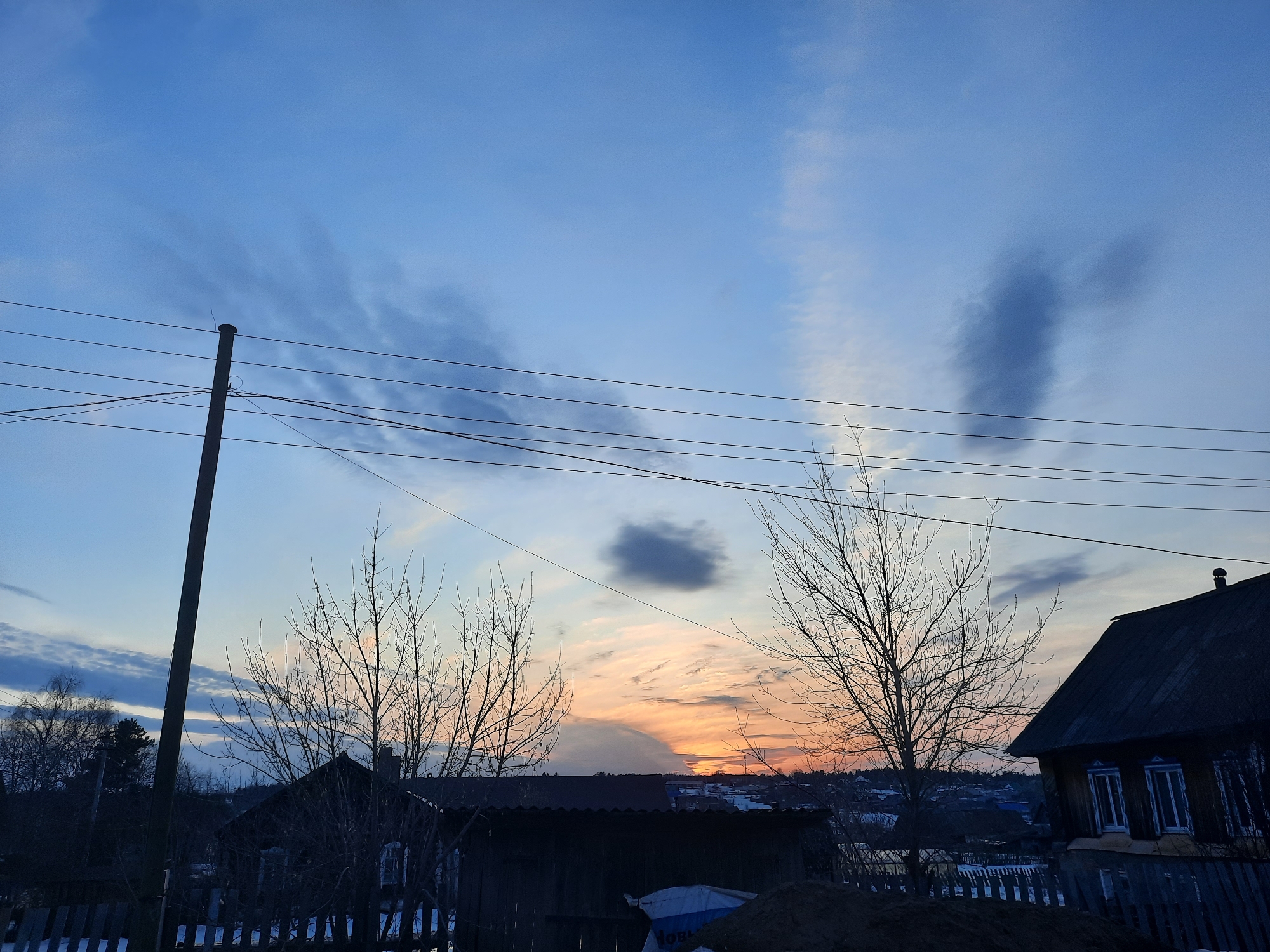 The image captures a rural or suburban scene at sunset, possibly in Krasnogorskoye, Russia, given the prompt. The sky transitions from blue to orange at the horizon, with scattered clouds. A utility pole stands on the left, with multiple overhead power lines stretching across the frame. Silhouetted against the sky are bare trees and the roofs of houses, with some fences visible at the bottom. The buildings appear to be older, with traditional architectural elements. There are no people visible in the image. The overall impression is one of a quiet, end-of-day setting in a residential area.