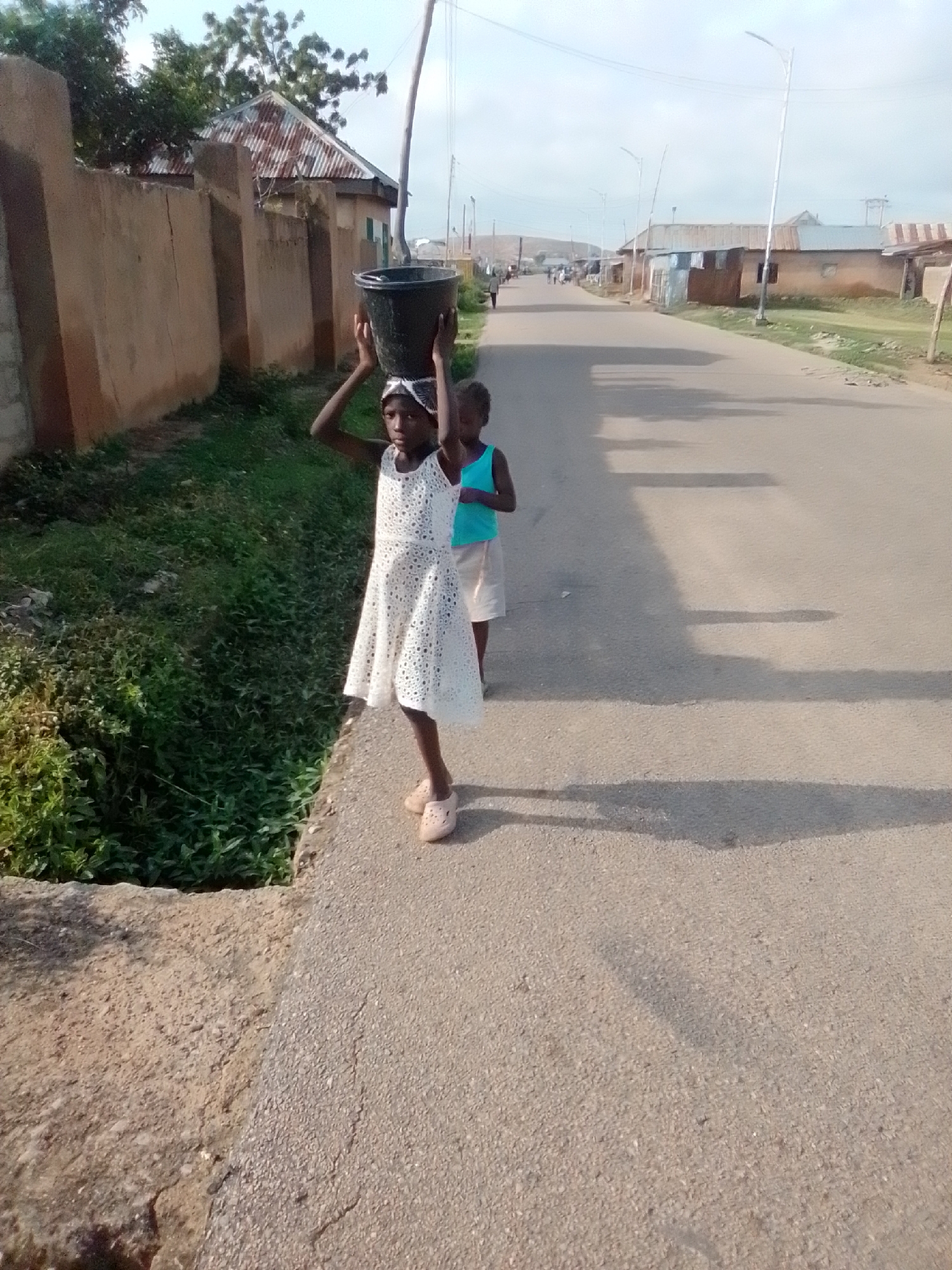 In the city of Gombe, Nigeria, two young girls are pictured walking on a paved road. The girl in the foreground, wearing a white dress, carries a black bucket on her head. Her companion, in a blue top and white shorts, walks slightly behind her. The environment features a wall on the left, overgrown with vegetation, and buildings with corrugated iron roofs lining the right side of the road. Streetlights are visible in the distance, and the sky is overcast.