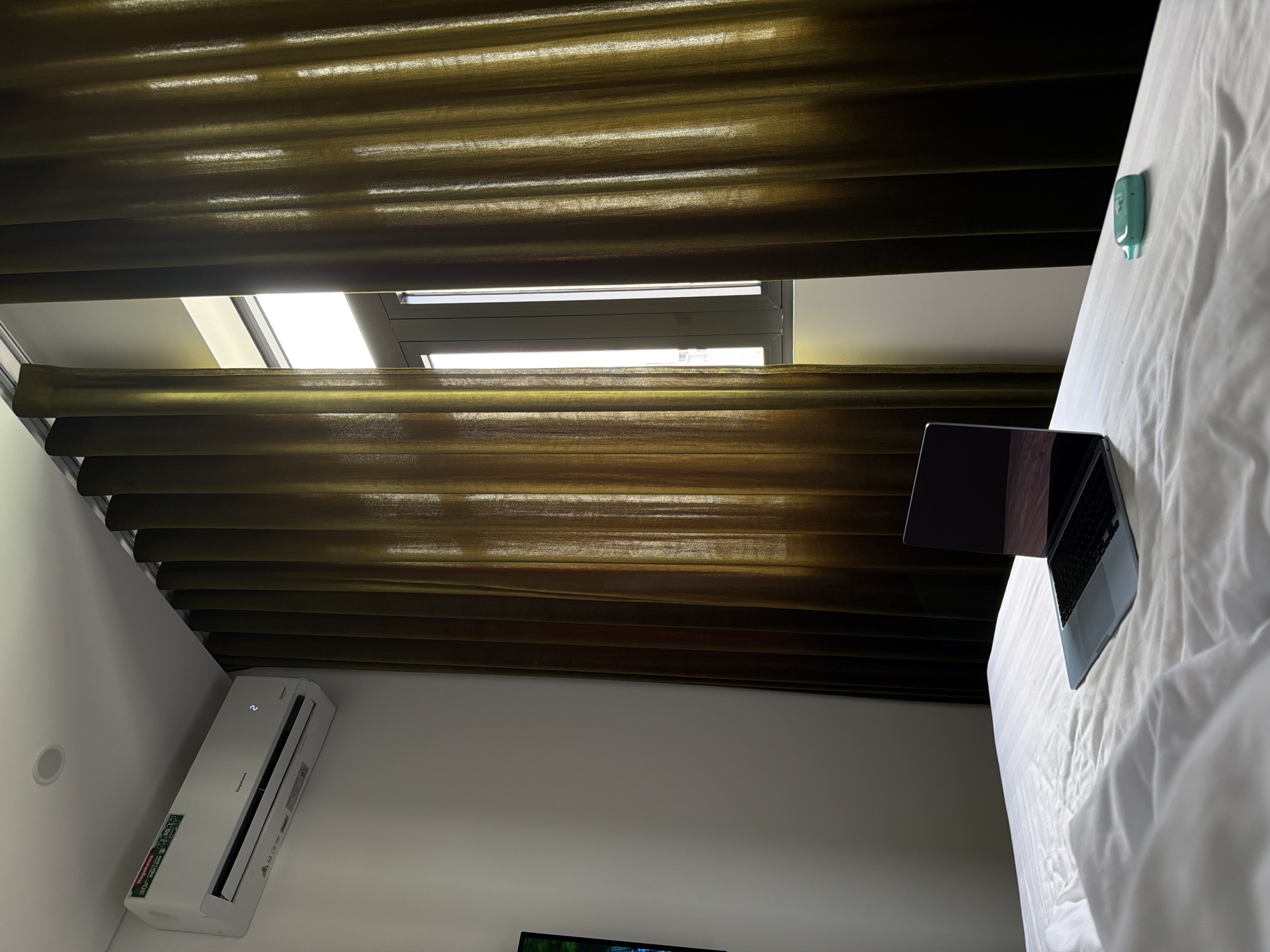 The image shows a room in what appears to be a hotel or residence in Đà Nẵng, Vietnam. The focus is on a bed covered with white linens, upon which a laptop is resting. The room has an air conditioning unit mounted on the wall, and window coverings appear to be partially closed, filtering sunlight. The view through the window coverings shows the presence of an exterior window frame, suggesting the room has access to outside light. The exact location is uncertain, but is likely within a building in the city of Đà Nẵng. No people are visible in the image.