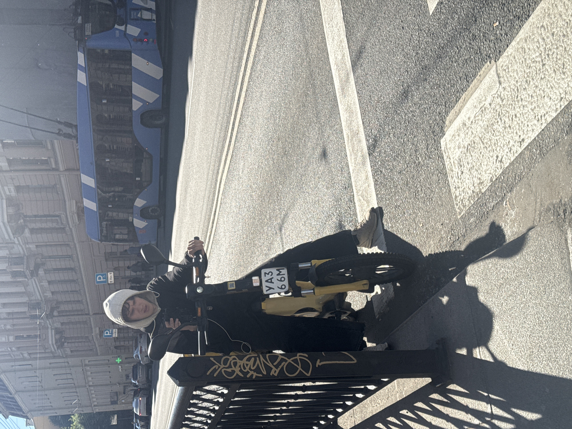 A person wearing a hooded sweatshirt and dark clothing is standing next to a yellow electric scooter on a city street in Saint Petersburg, Russia. The person is holding a smartphone and looking down at it. The scooter has a license plate that reads "YA3 66M". In the background, there is a blue and white striped bus, several cars, and buildings with neoclassical architecture. The street is paved with asphalt, and there are pedestrian crossings and double white lines. Sunlight casts long shadows on the ground. The scene appears to be captured during daylight hours.
