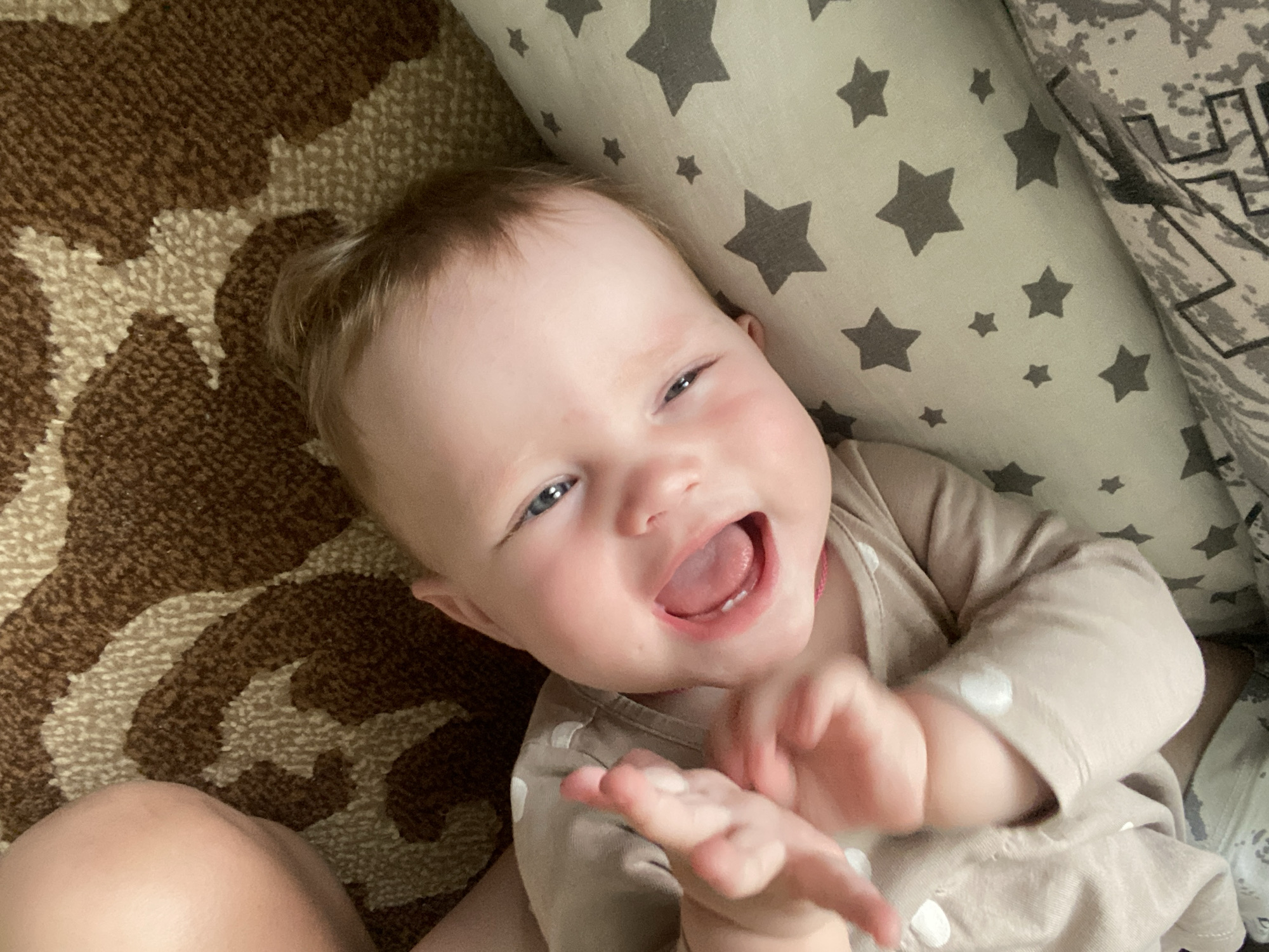 The image shows a close-up, overhead view of a baby lying down. The baby has fair skin, blue eyes, and light brown hair. The baby is wearing a light brown onesie with white polka dots. The baby's mouth is open in a wide smile, and its tongue is visible. The baby's hands are raised in front of its face, with fingers spread. The background consists of a carpet with a brown and beige pattern and a pillow with a grey star pattern. The image does not provide any information about the location in the city of Фоки, Russia.