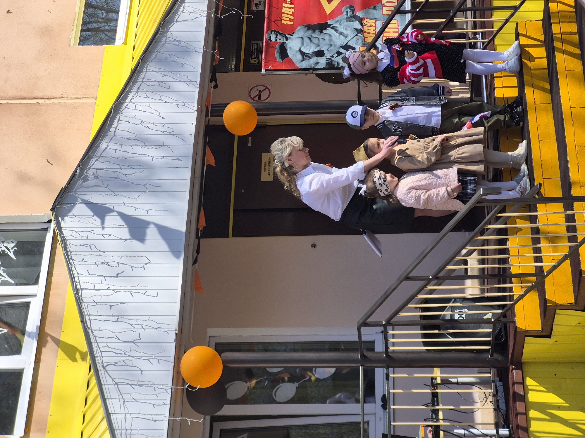 This outdoor scene shows an adult woman in a white blouse and black skirt standing on a set of stairs with three children. The woman is positioned so that she seems to be arranging or adjusting something on the children. The first child is wearing a pink coat and a patterned headband. The second child is wearing a beige dress, while the third child is in a jacket and a baseball cap. A fourth child, dressed in a black dotted dress with a white collar and cuffs, stands at the top of the stairs. All the children are wearing white tights.
The setting is decorated with orange balloons and bunting. A poster of a military figure is visible behind them, and a "No Smoking" sign. The building is painted in bright yellow and orange colors, with white trim. A window on the side has decorative paper cutouts. The scene is outside a building in Tobolsk, Russia.
