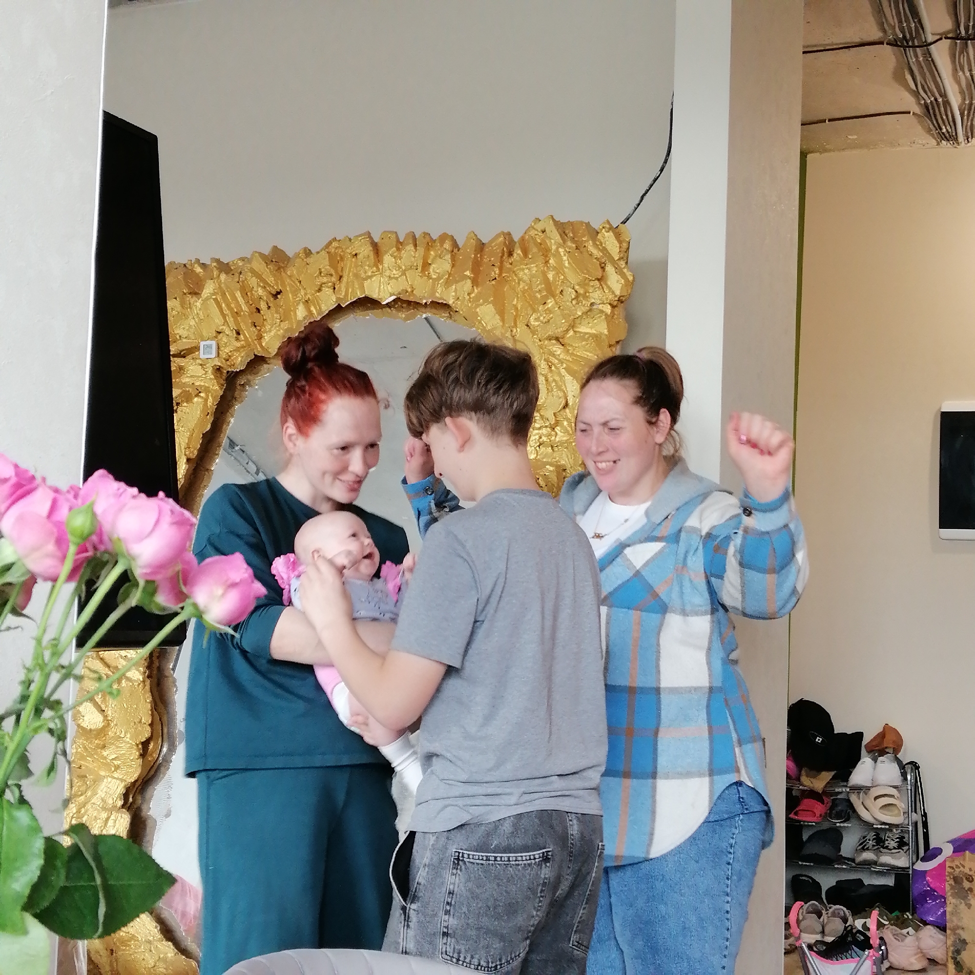 The image depicts a scene inside a domestic space, likely in Unknown, Unknown. Three individuals are present, a woman holding a baby, a young man in a grey t-shirt and grey jean shorts, and another woman in a blue plaid flannel shirt and jeans. The woman holding the baby has red hair pulled back in a bun and is wearing a dark green outfit, and the baby is wearing a pink onesie. The second woman has brown hair pulled back in a ponytail and is raising her fist in the air. They are standing in front of a large, ornate, gold-framed mirror. To the left of the frame are pink roses in a vase. The background includes a partially visible shoe rack with assorted shoes. There are unfinished walls with exposed electrical wires in the top right corner of the frame.