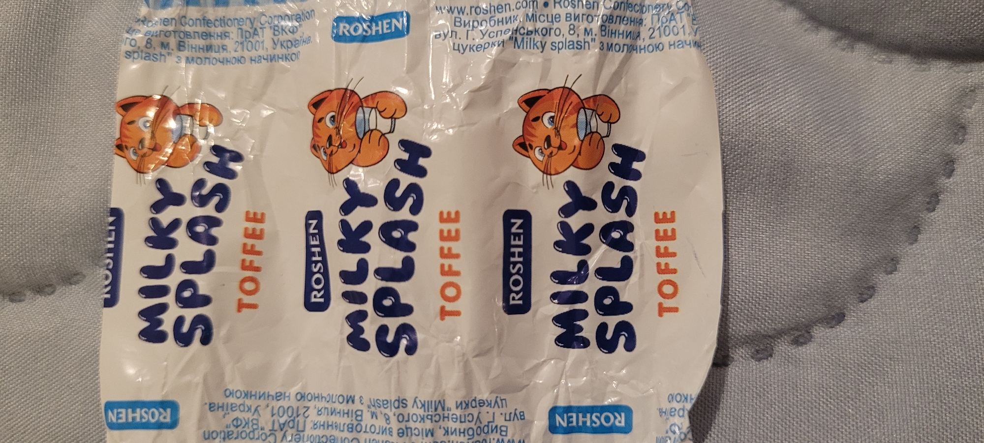 The scene is stated to be in Heusenstamm, Germany, although the image provides no visual indicators of this specific geographic location. The primary subject is a section of a white candy wrapper, which is repeated three times vertically, suggesting either a continuous roll of packaging or multiple individual wrappers. The wrapper features the brand name "ROSHEM" in a dark blue rectangular box and the product name "MILKY SPLASH" in large, bubbly dark blue capital letters. Below this, the word "TOFFEE" is printed in orange capital letters. Adjacent to the text, a cartoon illustration of an orange cat with stripes, a smiling expression, and blue eyes is depicted holding a white cup containing a light blue liquid.
Along the left edge of the wrapper, vertical text in Ukrainian is visible, identifying the manufacturer as "ROSHEM Confectionery Corporation PrAT "VKF"" and stating the manufacturing location as "Вінниця, 21001, Україна" (Vinnytsia, 21001, Ukraine). The text also describes the product as "цукерки "Milky splash" з молочною начинкою" (candy "Milky splash" with milk filling). Similar, partially obscured text is present along the right edge, rotated 180 degrees. The wrapper material appears to be flexible plastic or foil, showing multiple creases and reflections from ambient light. The wrapper rests on a light grey fabric surface, which exhibits a woven texture and subtle, regularly spaced dot-like indentations or patterns.