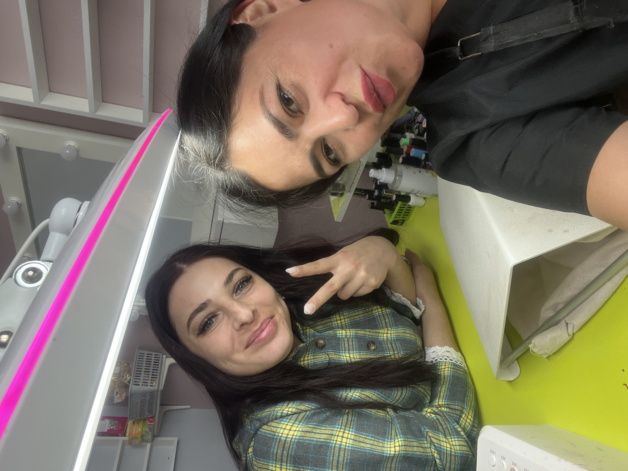 This image captures two women posing for a photo in what appears to be a beauty or nail salon, likely in Chaikovsky, Russia. The image is oriented with the women's heads to the left.
The woman in the upper portion of the frame has dark hair pulled back, prominent dark eyebrows, and is wearing dark lipstick. She looks directly at the camera with a slightly pensive or stylish expression, her lips slightly parted. She is dressed in a dark-colored top or apron with a visible strap and buckle.
The second woman, positioned below and slightly behind the first, has long dark hair and blue eyes. She is smiling gently and making a peace sign with her right hand. She wears a green, yellow, and dark grey plaid shirt with white lace detailing on the cuffs.
The setting features a bright, professional environment. A prominent white lamp with a vibrant pink stripe runs diagonally across the left side of the frame, suggesting specialized lighting for beauty procedures. In the background, there's a vanity mirror with globe-style bulbs. Various small bottles, likely nail polishes or cosmetics, are visible on shelves behind the first woman. A white spray bottle and a device with visible text "30S," "60S," "120S" (indicating a timer, possibly for a nail lamp) are also present. The surface supporting the lower woman appears to be a bright lime green, contrasting with the light pink wall behind her. The overall mood is casual and friendly, as the women are clearly posing for the camera.