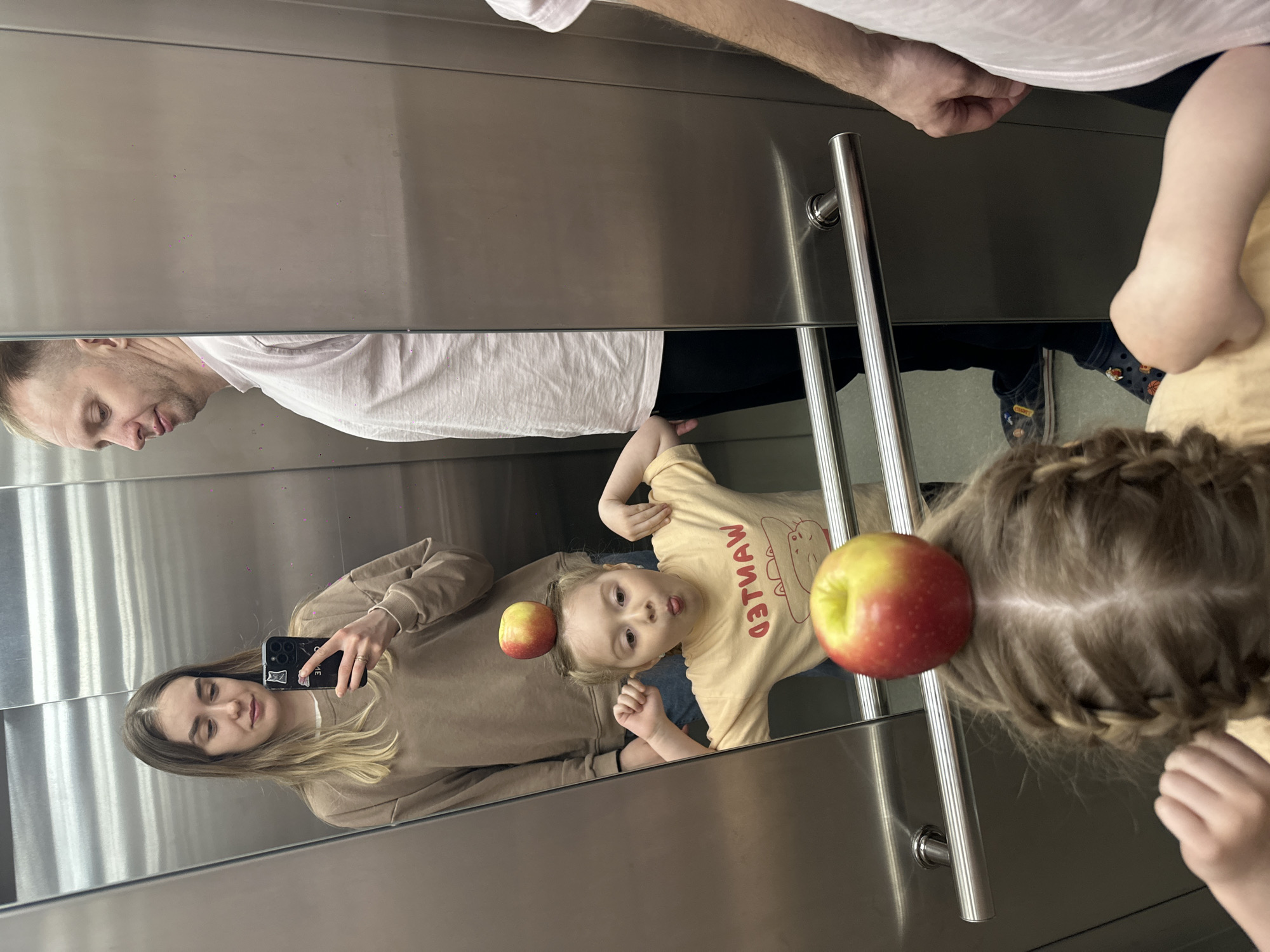 The photograph, taken in what appears to be an elevator, depicts a family. The reflection in the elevator's mirrored wall shows a woman holding a smartphone as if taking a picture. A girl in a yellow shirt with the word "WANTED" on it poses. There are two apples, one on the girl's head, and another on the head of a second girl whose braided hair is visible. A man in a white shirt is partially visible at the top of the mirror. The location is potentially Glazov, Russia, based on external information.