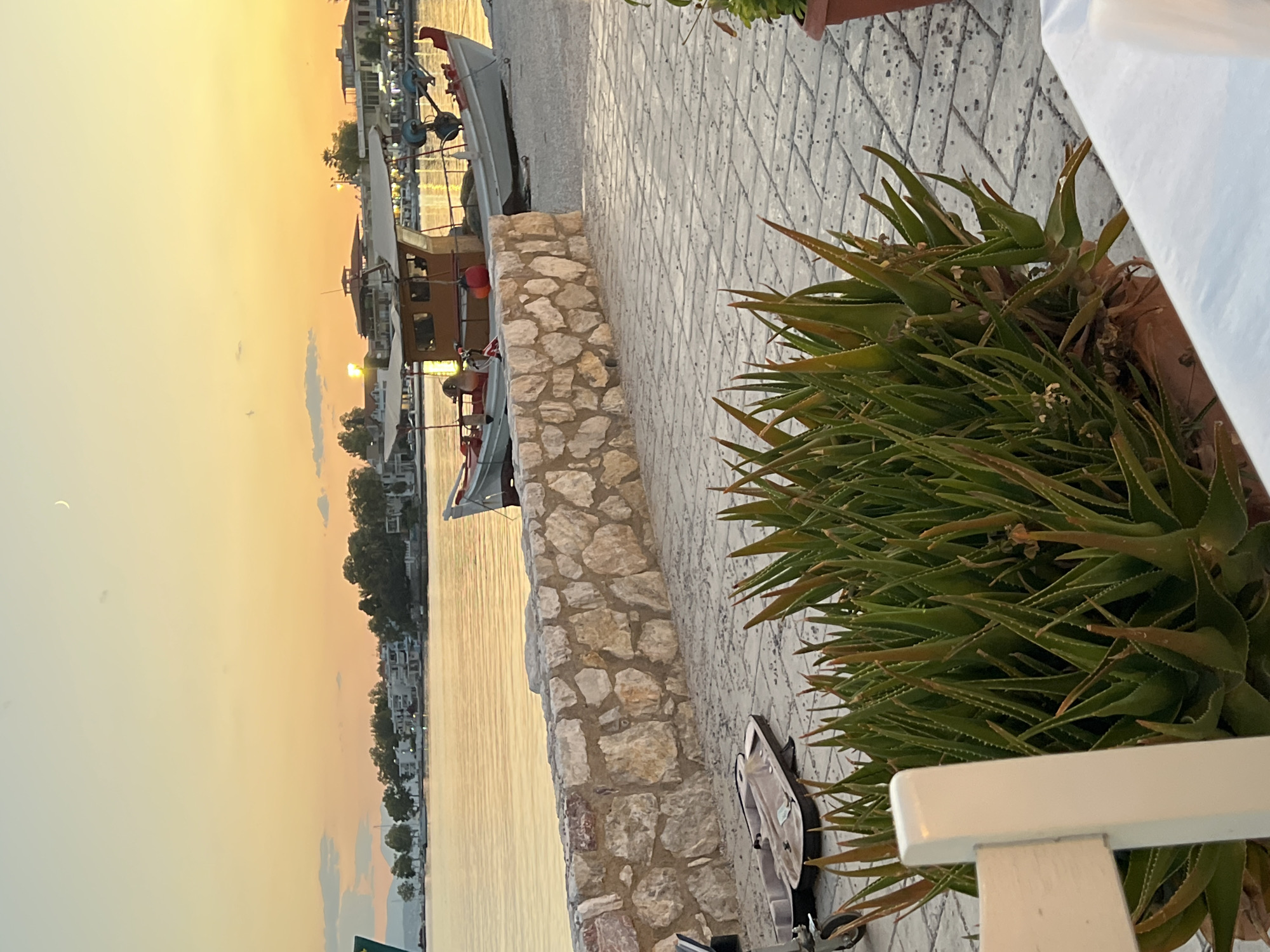 A wide shot captures a serene waterfront scene at dusk in Eretria, Greece. The sky is painted with soft hues of orange and yellow, with a crescent moon visible. A calm body of water reflects the warm light, and in the distance, a line of buildings and trees forms the coastline.
In the foreground, a paved walkway is bordered by a low, rough-hewn stone wall. A large pot of vibrant green aloe vera plants sits on the walkway, with spiky leaves reaching upwards. To the left of the aloe, a white chair is partially visible.
On the walkway, a light-colored case, possibly for a musical instrument, is open. Further back, a fishing boat with a wooden cabin rests on the shore, its hull painted white with red accents. The boat is equipped with fishing gear, including nets and lines.
The overall impression is one of peaceful tranquility, with the natural beauty of the sunset and the gentle lapping of the water creating a soothing atmosphere.