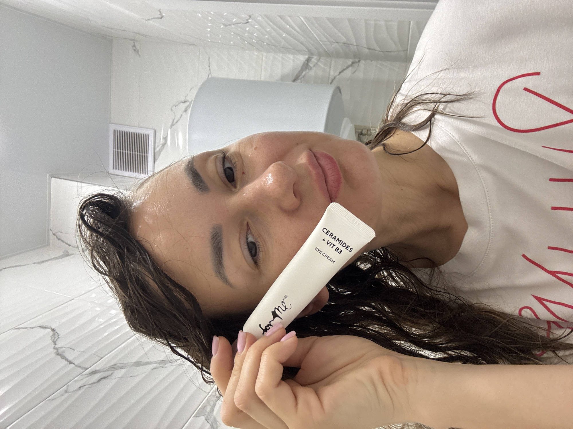The image shows a woman lying on her side, holding a tube of "for me" brand eye cream, specifically "CERAMIDES + VIT B3". She appears to be in a bathroom, evident from the tiled floor and walls, a wall-mounted toilet tank, and a ventilation grill. The woman has wet hair, and her skin appears slightly dewy. The image does not depict any specific location in the city of Sochi, Russia.