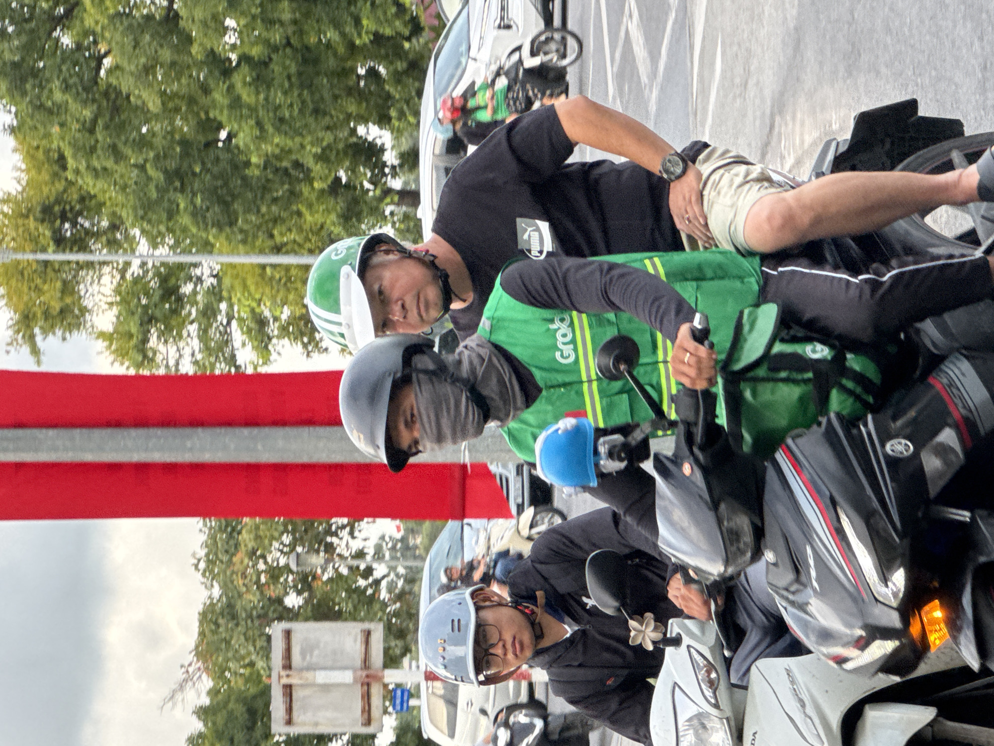 In Thuận Hóa Ward, Vietnam, three individuals are observed on two motorcycles, engaged in travel. The foremost motorcycle, a dark-colored model, carries a young male driver wearing a gray helmet and eyeglasses, alongside a passenger obscured by a green vest and a black face covering. Their motorcycle is white. The second motorcycle is ridden by a man with a green and white helmet, wearing a black t-shirt with a Puma logo and light-colored shorts. He is seen looking to his left, with his right hand on the handlebars. A third individual on a separate motorcycle, wearing a red helmet, is partially visible in the background. The scene is set against a backdrop of lush green trees and a blurred red banner, suggesting an urban environment. The lighting indicates it is daytime, with an overcast sky.