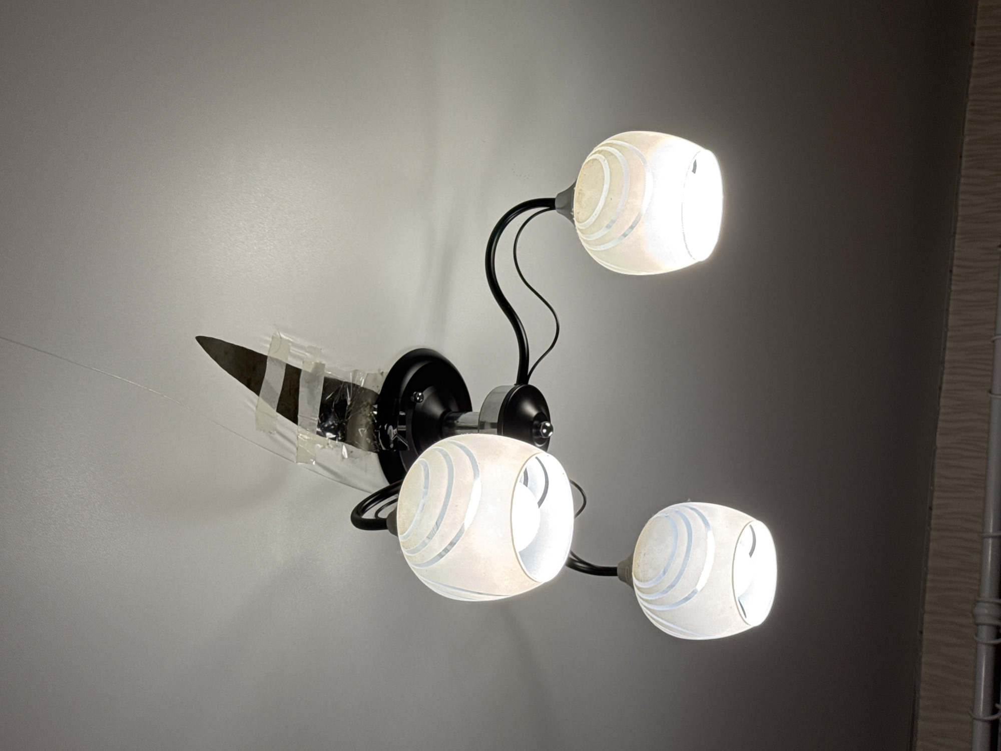 A three-bulb ceiling light fixture with white, frosted glass shades is suspended from a white ceiling. The shades have a swirling blue pattern. The fixture is black metal. The central part of the fixture appears to be taped with clear packing tape and a dark piece of material is attached. The lighting suggests an indoor setting, possibly a residential room in Kondratovo, Russia. The bottom of the image shows the edge of a patterned wallpaper and a pipe or conduit.