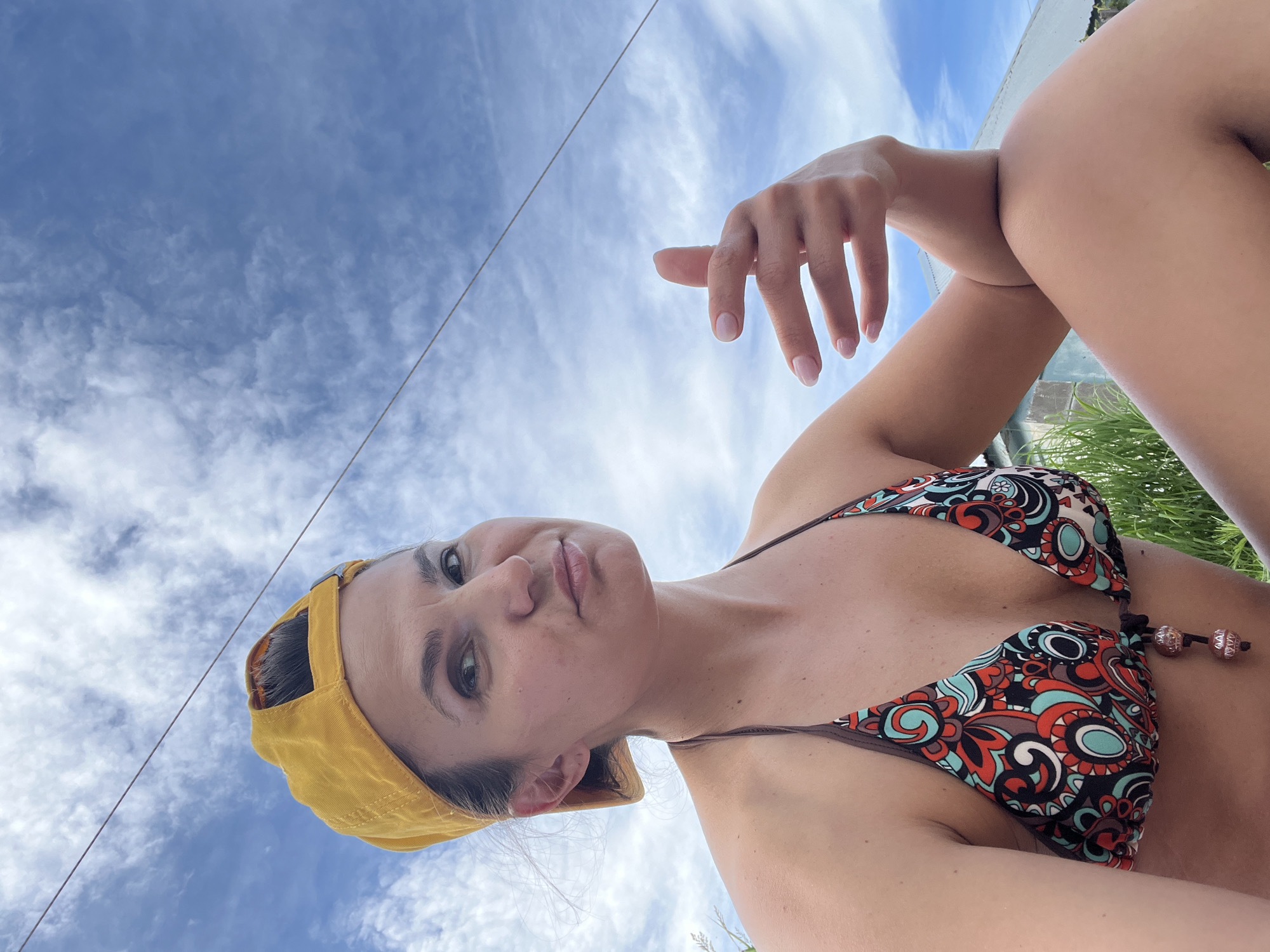 A woman wearing a yellow baseball cap and a colorful bikini top is pictured outdoors. She is looking towards the camera with her right hand raised, fingers spread and extended. The background consists of a blue sky with scattered white clouds and a faint wire running across the upper portion of the frame. Some greenery and a portion of a building are visible in the lower right of the image. The lighting suggests it is daytime. The location is not specified as Petropavlovsk-Kamchatsky, Russia.