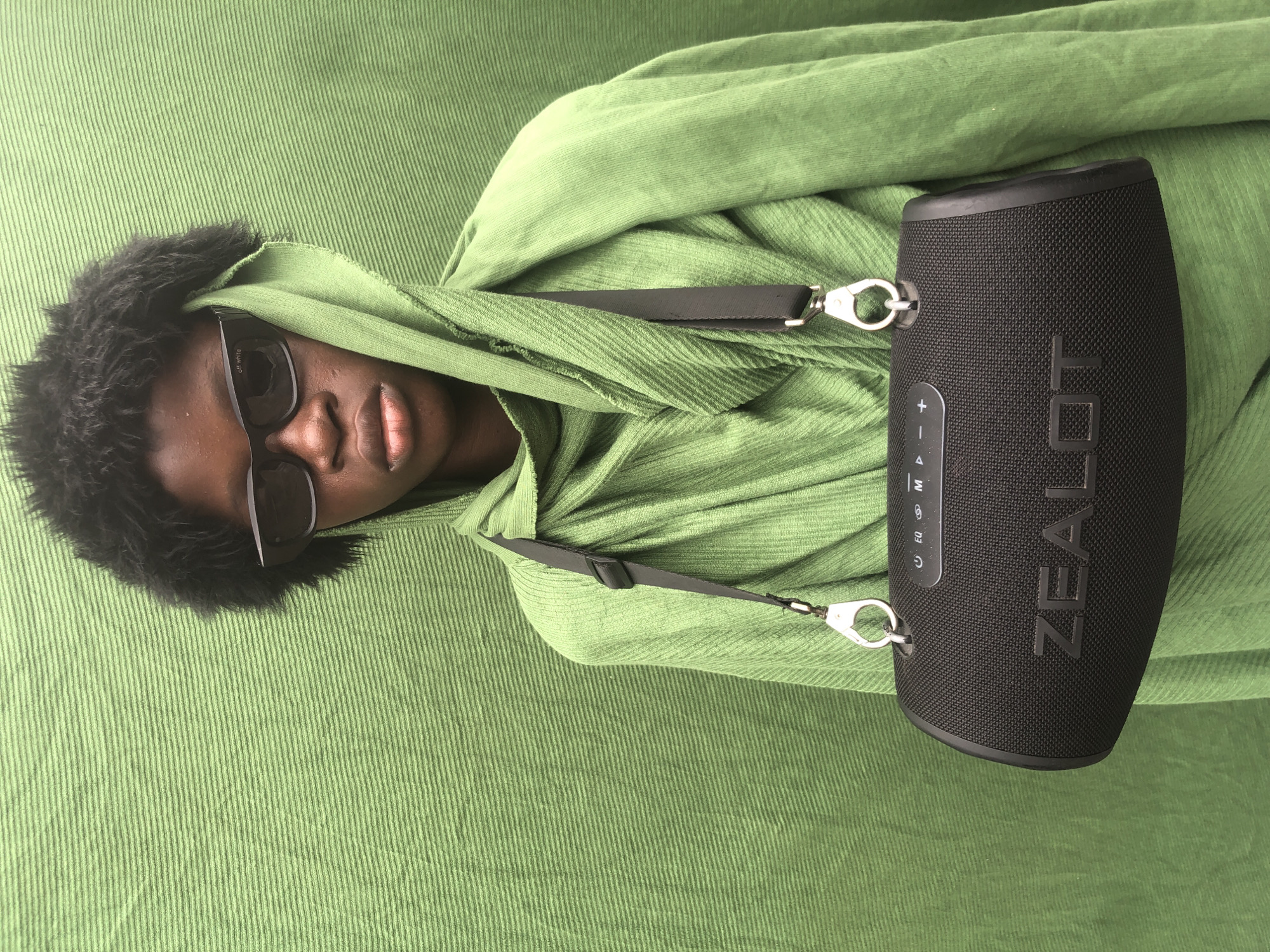A young person stands against a textured green backdrop in Ado, Nigeria. They have dark, textured hair and are wearing black sunglasses. A green, draped garment covers their head and shoulders. A black portable speaker with the brand name "ZEALOT" visible is slung across their chest with a strap. The speaker has control buttons visible on its surface. The lighting is even, suggesting an outdoor or well-lit indoor setting.