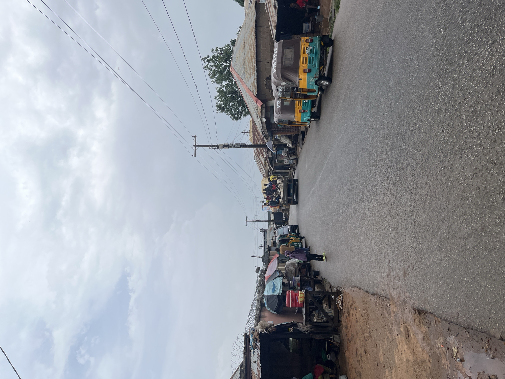 The image depicts a street scene in Jos, Nigeria. The street is paved and appears wet. A row of buildings with corrugated metal roofs lines one side of the street, interspersed with informal stalls or shops. Wires for power lines run overhead. A number of auto rickshaws (tuk-tuks) are parked along the street. People can be seen walking or gathering in small groups near the vehicles and stalls. The sky is overcast. The scene has a generally bustling, informal atmosphere.
