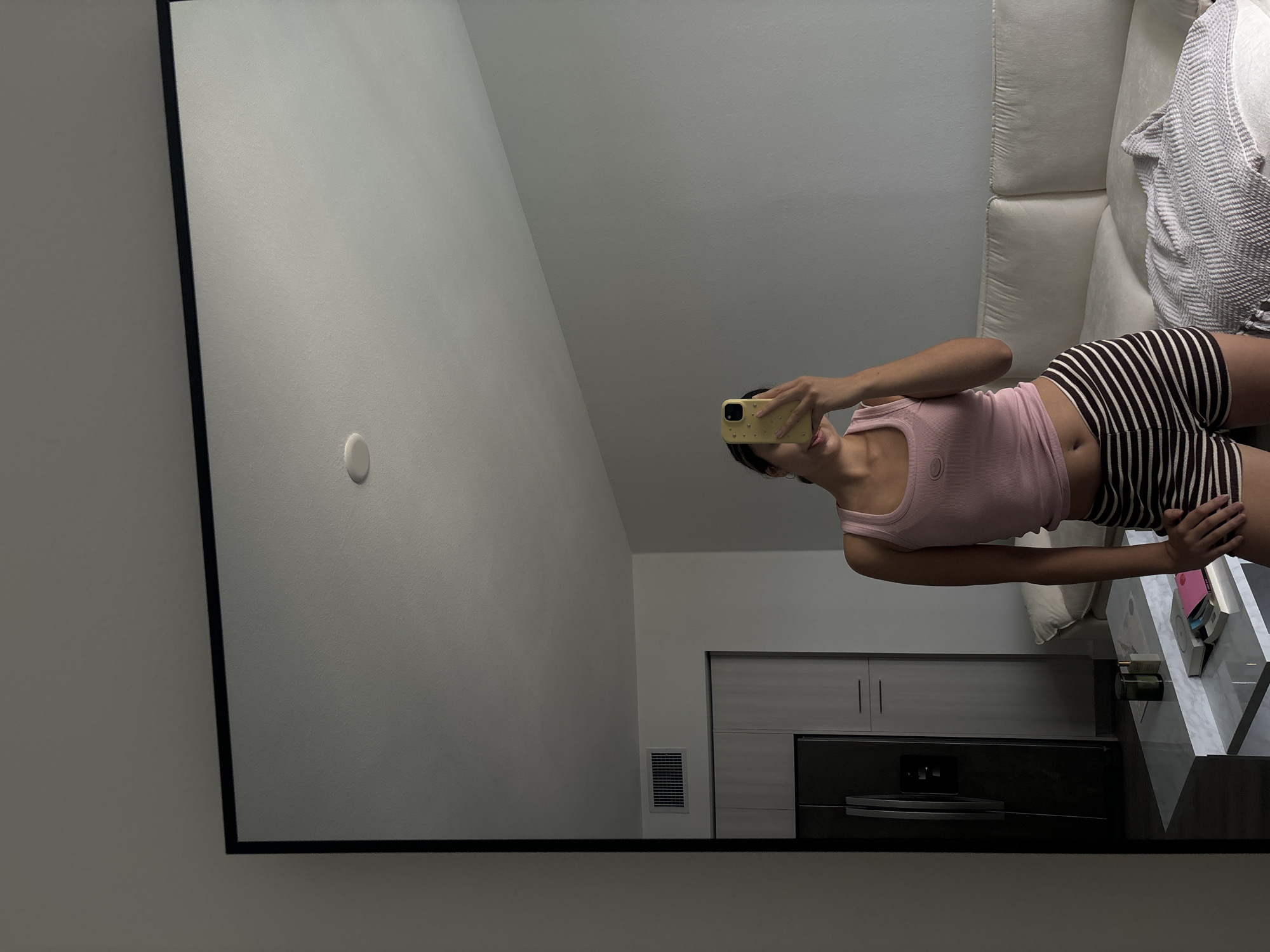A photograph captures a reflection in a mirror, presenting a woman taking a selfie. She is dressed in a light pink tank top and striped shorts. The scene reflected in the mirror includes a portion of a white sofa with a patterned throw, a modern coffee table with items on it, a kitchen area with cabinets and appliances, and a wall with a vent. The location appears to be an interior space in Boca Raton, United States of America.