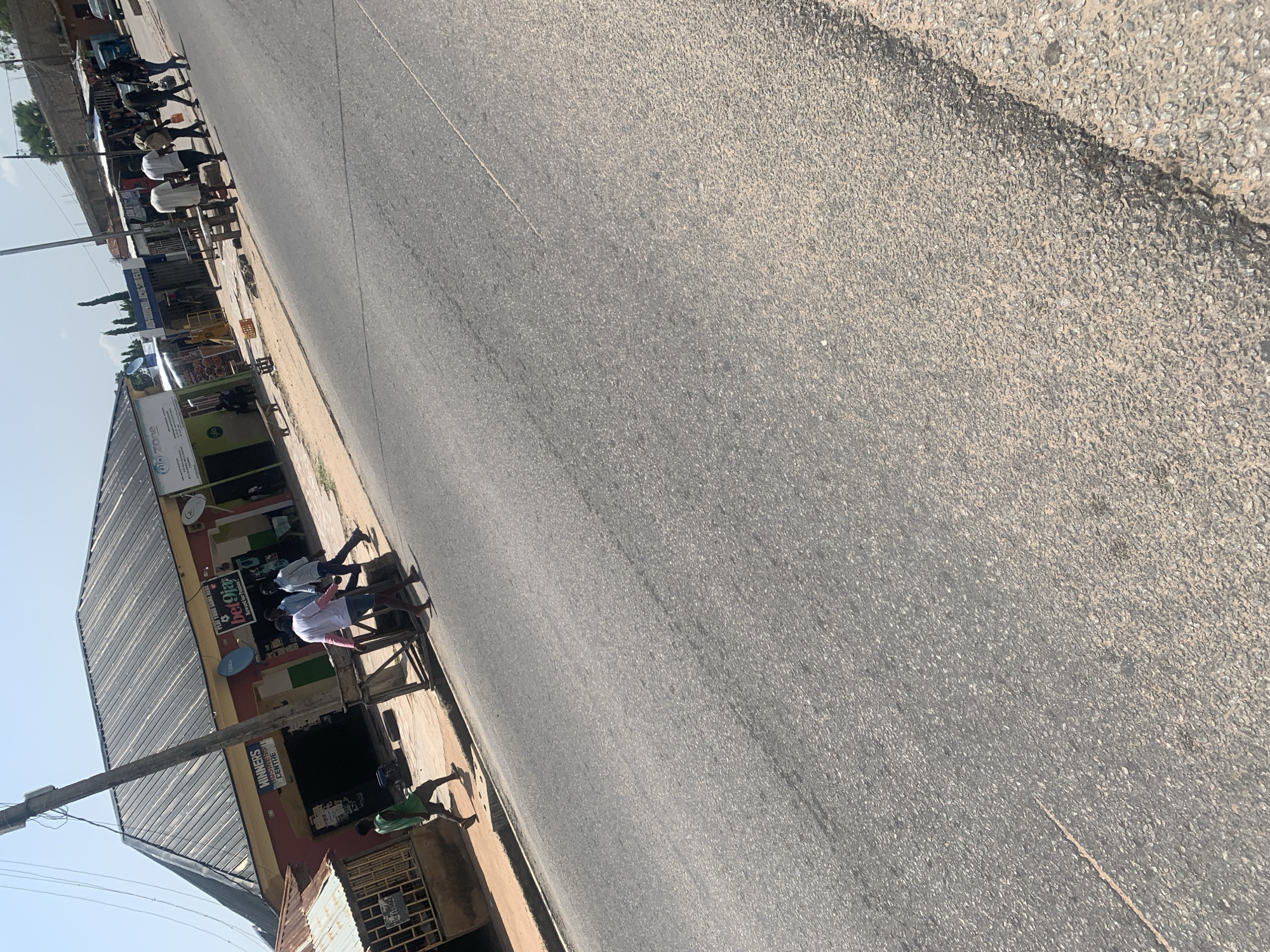 The image shows a street scene in Jalingo, Nigeria. On the left, a man in a green shirt walks on the sidewalk in front of a building with a corrugated metal roof. To the right of this building, there is a larger building with a dark gray corrugated metal roof, painted red and yellow. This building has signage that includes "WINNERS BUSINESS CENTRE" and "bet9ja." Several people are walking on the sidewalk in front of this building and crossing the street. One man in a pink shirt is facing away from the camera, and two men in light-colored shirts are walking in the same direction. Further to the right, a group of people are crossing the street from right to left. The street itself is paved with asphalt and appears to be in moderate condition. The environment is sunny with a clear blue sky. The overall scene depicts a typical day in a commercial area of the city.