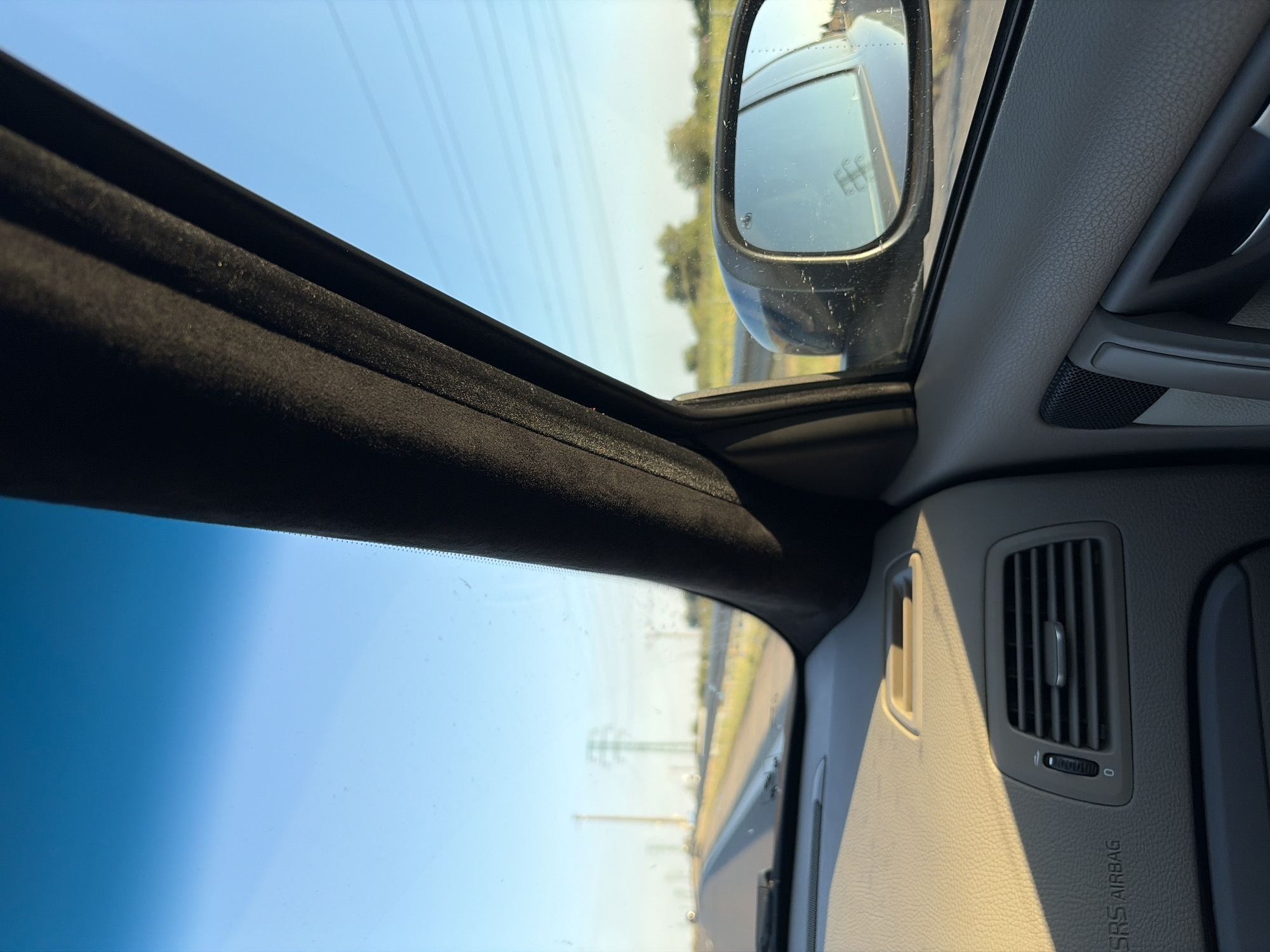The image shows the interior of a car looking out the passenger side window. A windshield pillar dominates the right side of the frame, with the blue sky and a distant horizon visible beyond it. The dashboard is visible in the foreground, with an air vent and the "SRS AIRBAG" warning label. In the side mirror, a reflection of the road and overhead power lines can be seen. The scene suggests a car journey, possibly on a highway, given the presence of power lines and the general openness of the environment. The provided context places the image in Vlčkovce, Slovakia, a rural area in Slovakia.