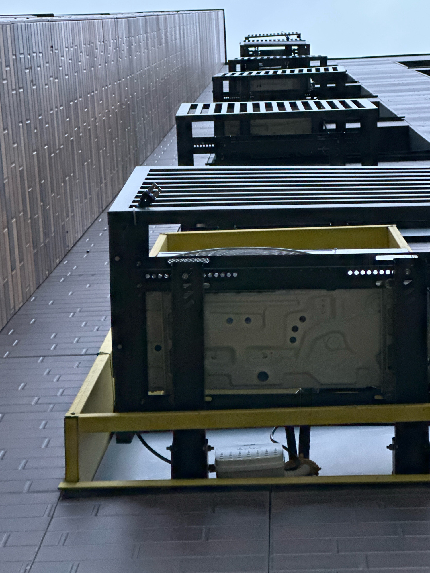 The image shows an upward-angled shot of an external air conditioning unit mounted on the side of a modern building. The building's facade is clad in dark, textured panels. Above the air conditioning unit, there are multiple levels of black metal grating or platforms, suggesting a fire escape or external walkway structure that ascends the building. The unit itself is housed within a black metal frame and sits on a bright yellow support structure. Various wires and components are visible beneath the unit, including a white electrical box. The sky is overcast, contributing to the muted lighting. The image does not provide any specific visual cues to place it in the city of Vidnoye, Russia.
