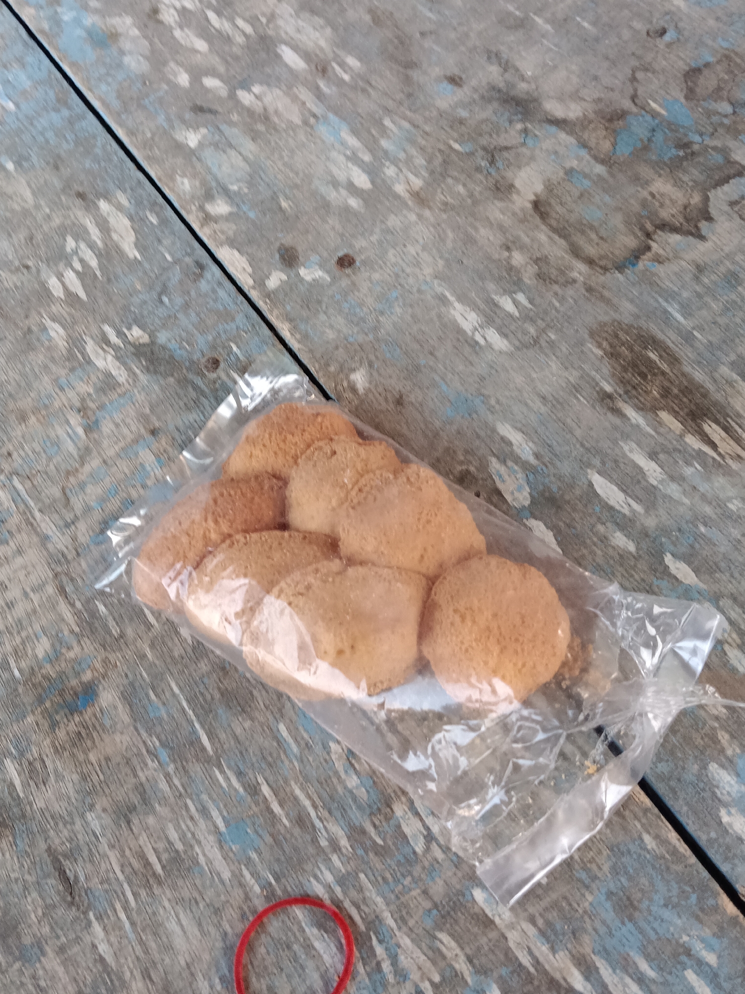 A package of round, golden-brown cookies sits on a weathered, blue-painted wooden surface. The cookies are arranged in two rows, with some overlapping, inside a clear plastic bag. The wooden surface shows signs of wear and tear, with visible grain and chipped paint. A red rubber band is partially visible in the bottom left corner of the image. The lighting is natural, casting soft shadows on the cookies. This scene is not specifically located in Tegal Gondo, Indonesia, but rather depicts a generic setting.