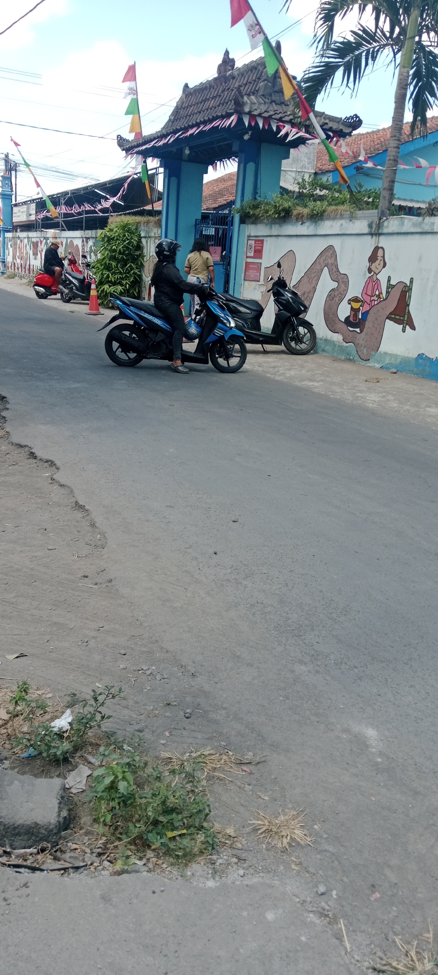 The image depicts a street scene in Purwomartani, Indonesia, near what appears to be a school or community building. A blue gate with decorative roofing and flags is visible, leading to the building. A mural of a cartoon-like character is painted on the wall beside the gate. Several motorbikes are parked along the roadside. In the foreground, two individuals are on motorbikes, one wearing a black helmet and jacket, and the other a yellow shirt. Another person is seated on a red motorbike further down the street. The road is paved with asphalt, and there is some vegetation and debris along the side. The sky is partly cloudy.
