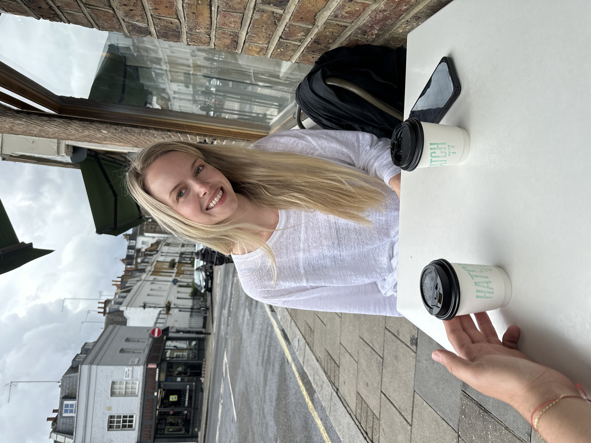 A blonde woman is sitting at a table outside a cafe, smiling at the camera. She is wearing a white long-sleeved shirt. In front of her are two disposable coffee cups, one with a black lid and the other with a black lid and a straw, and a smartphone. A person's hand is visible on the left side of the frame, reaching towards one of the coffee cups. Behind the woman, a street scene is visible with buildings and cars. The street appears to be wet, suggesting it may have recently rained. The cafe appears to be located in London, United Kingdom, as indicated by the architecture of the buildings and the presence of a red traffic sign.