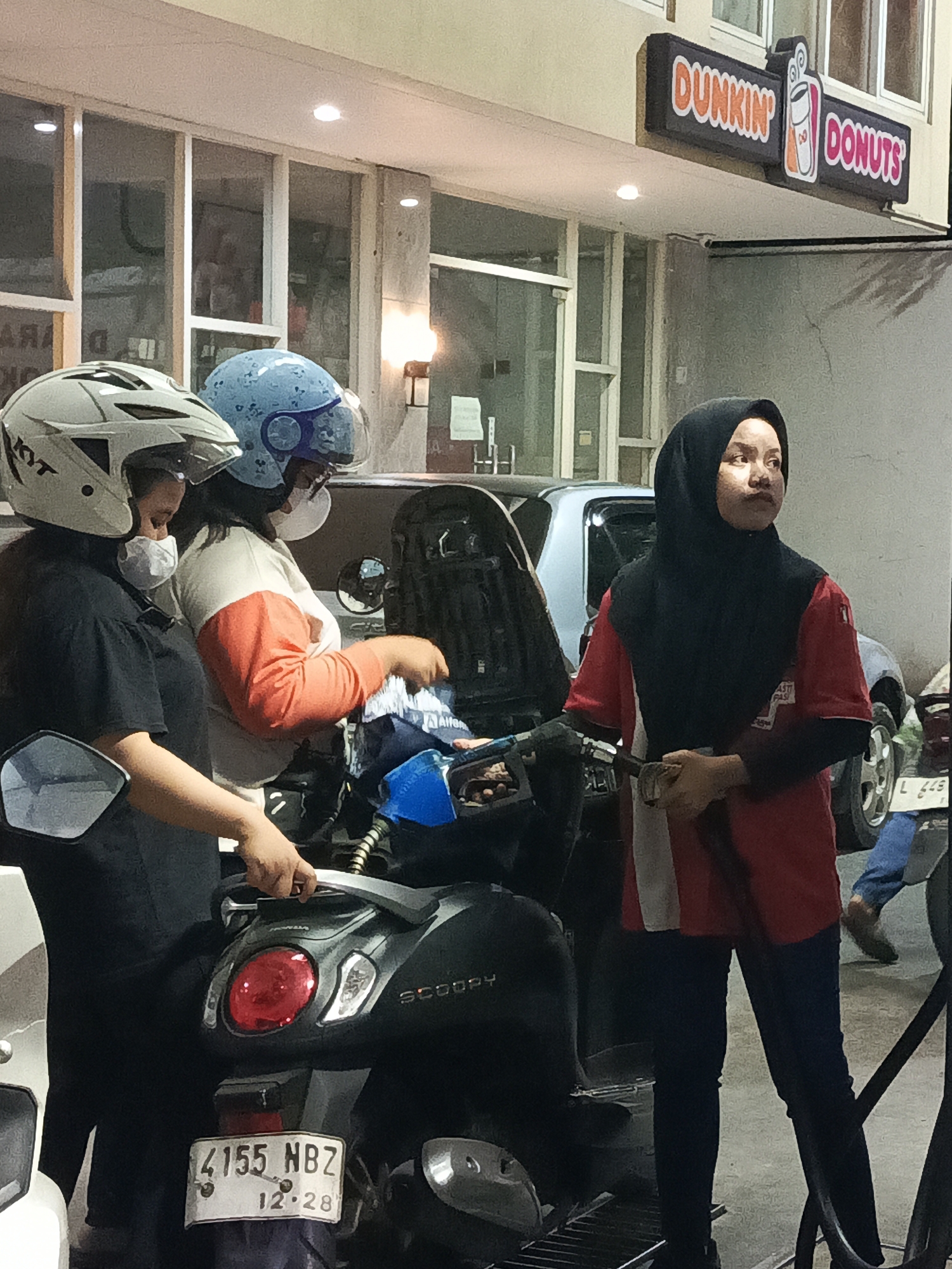 In Surabaya, Indonesia, at a Dunkin' Donuts establishment, two individuals wearing helmets and masks are near a scooter. One person, wearing a white helmet and a black shirt, stands on the left side of the scooter. To their right, another person in a blue helmet and an orange and white shirt is attending to the scooter's fuel tank with a blue nozzle. Further to the right, a woman in a red shirt and a black hijab is holding a fueling nozzle. A car is visible in the background, and the Dunkin' Donuts sign is prominently displayed. The scene appears to be at a gas station with a Dunkin' Donuts attached.