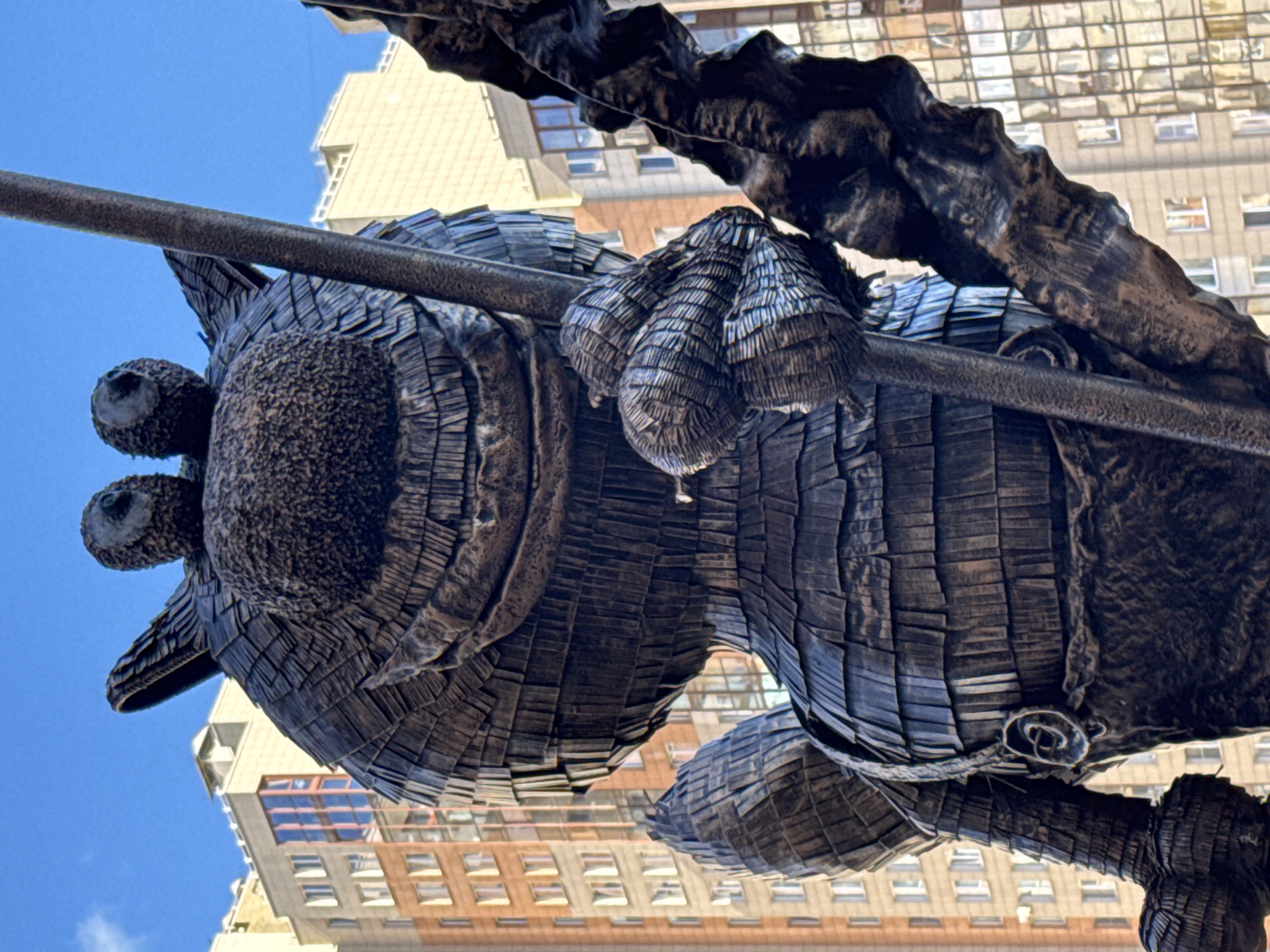 This image shows a low-angle view of a bronze sculpture of a cat, likely a public art installation, against a backdrop of buildings and a blue sky. The cat is depicted with stylized, textured surfaces, giving it a segmented, almost wooden appearance. It appears to be holding onto a horizontal pole or branch. Buildings with numerous windows are visible in the background, indicating an urban environment. The location is likely Saint Petersburg, Russia, given the architectural style of the buildings and the presence of similar public art.