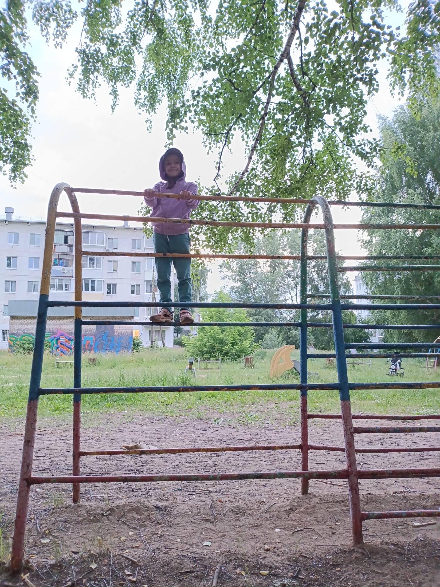 A young girl wearing a purple hooded sweatshirt and green pants stands on a horizontal bar of a metal playground structure. The structure is colored with rust-colored supports and blue and green bars. In the background, there is a multi-story apartment building with balconies and windows. The area around the playground is covered in grass and dirt. Trees with green leaves are visible above and behind the playground. The sky is overcast. The image was taken in the city of Chaikovsky, Russia.