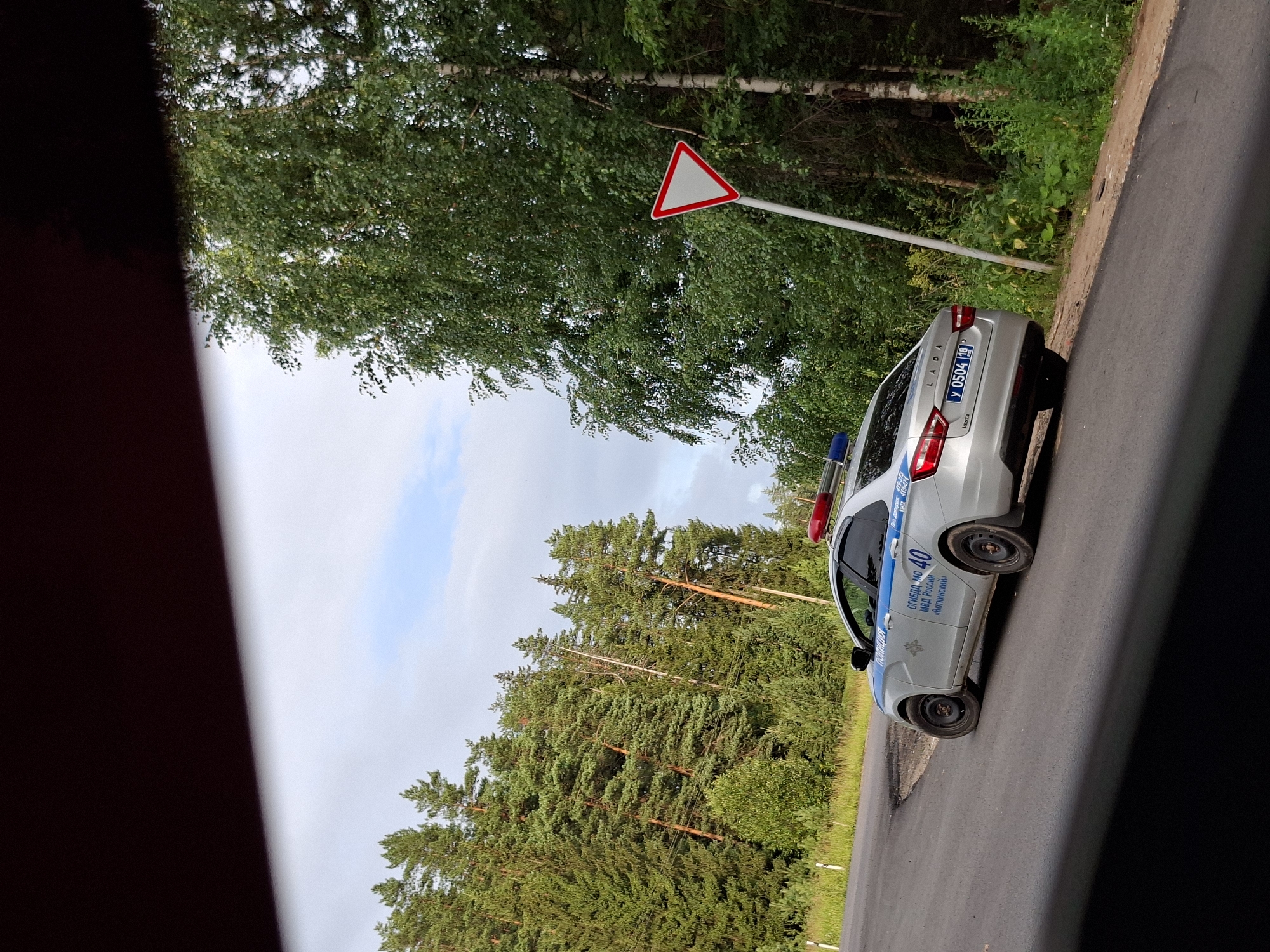 A silver Lada Vesta police car with blue and red emergency lights on its roof is parked on the side of a paved road that curves to the left. The car's markings include "ПОЛИЦИЯ" (Police) in blue lettering, a circular police emblem, and the text "ОГИБДД МО МВД РОССИИ «Вояткинский»" (Department of State Road Safety of the Ministry of Internal Affairs of Russia "Votkinsk District"). The license plate reads "У 0504 18". The car is positioned on the right side of the road, facing away from the viewer.
The road is surrounded by a dense forest of tall green trees, including pines and birch trees. A red and white triangular yield sign is mounted on a pole to the right of the road. The sky is overcast with some patches of blue. The foreground is partially obscured by a dark, blurry object, possibly a vehicle or a shadow. The scene appears to be in a rural or semi-rural area, consistent with the Votkinsk District in Russia. No people are visible in the image.