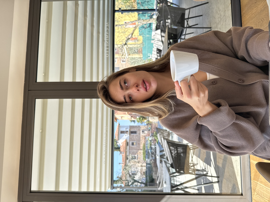 A young adult female is depicted seated indoors at what appears to be a restaurant or cafe, specifically identified as "Il Caminetto" in Albisano, Italy. She has light brown hair and is wearing a brown ribbed cardigan over a white top. Her right hand holds a white ceramic mug. She looks directly forward with slightly parted lips. Behind her is a large glass partition revealing an outdoor terrace with numerous unoccupied dark-colored tables and chairs. Beyond the terrace, a clear view of Albisano's urban landscape under bright daylight is visible, featuring traditional Italian buildings with red-tiled roofs, light-colored facades, and scattered trees. Reflected on the upper part of the glass are horizontal window coverings and the inverted text "IL CAMINETTO".