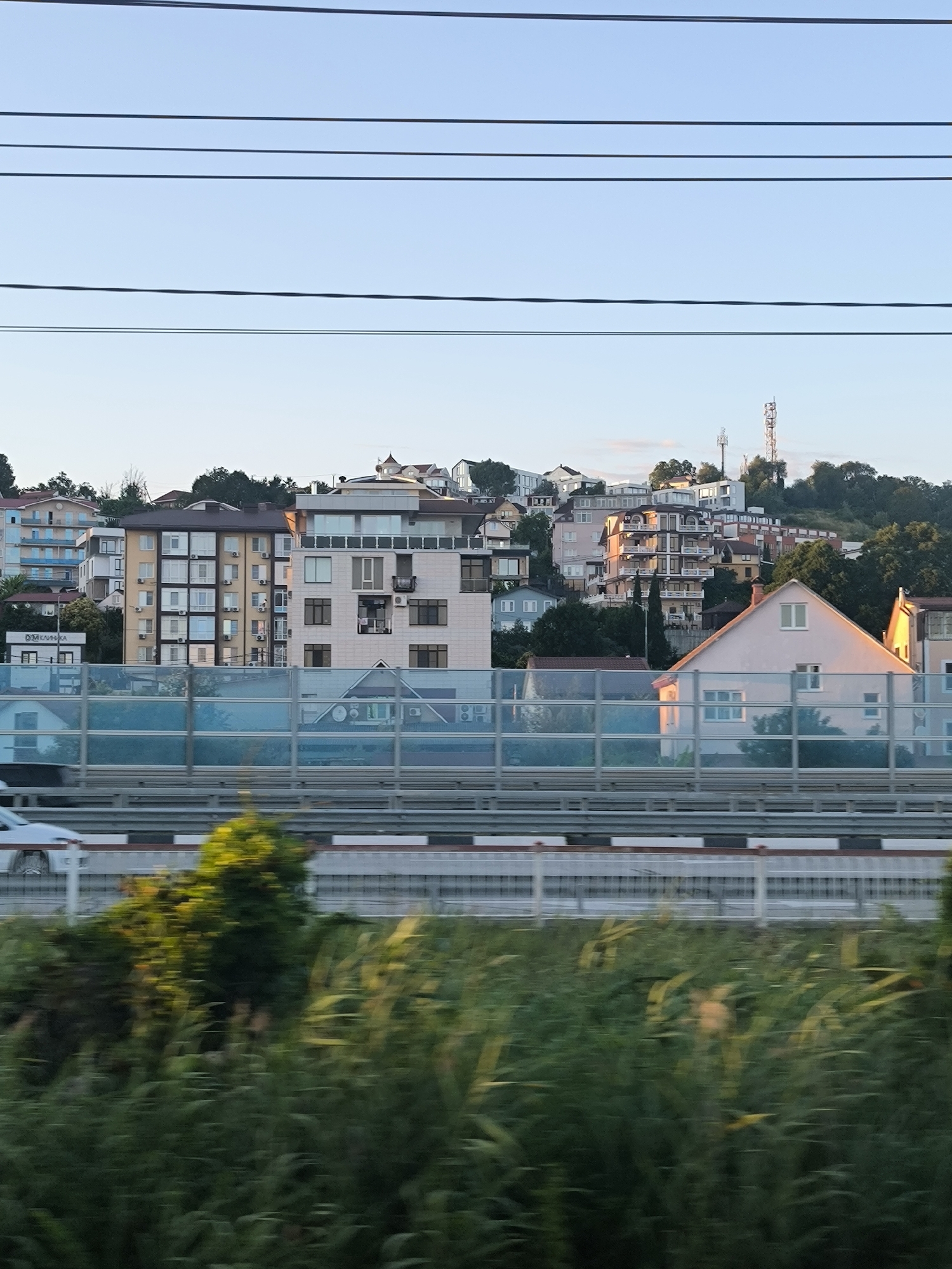 A high-angle, medium shot captures a cityscape with numerous residential buildings and infrastructure. The buildings are of various heights and architectural styles, situated on a hillside under a clear sky. Power lines are visible across the top of the frame. A highway with a sound barrier runs horizontally through the middle ground. A white car is partially visible on the highway, suggesting movement. In the foreground, blurred green vegetation indicates the photo was taken from a moving vehicle, possibly a train or car. The lighting suggests it is either early morning or late afternoon.
