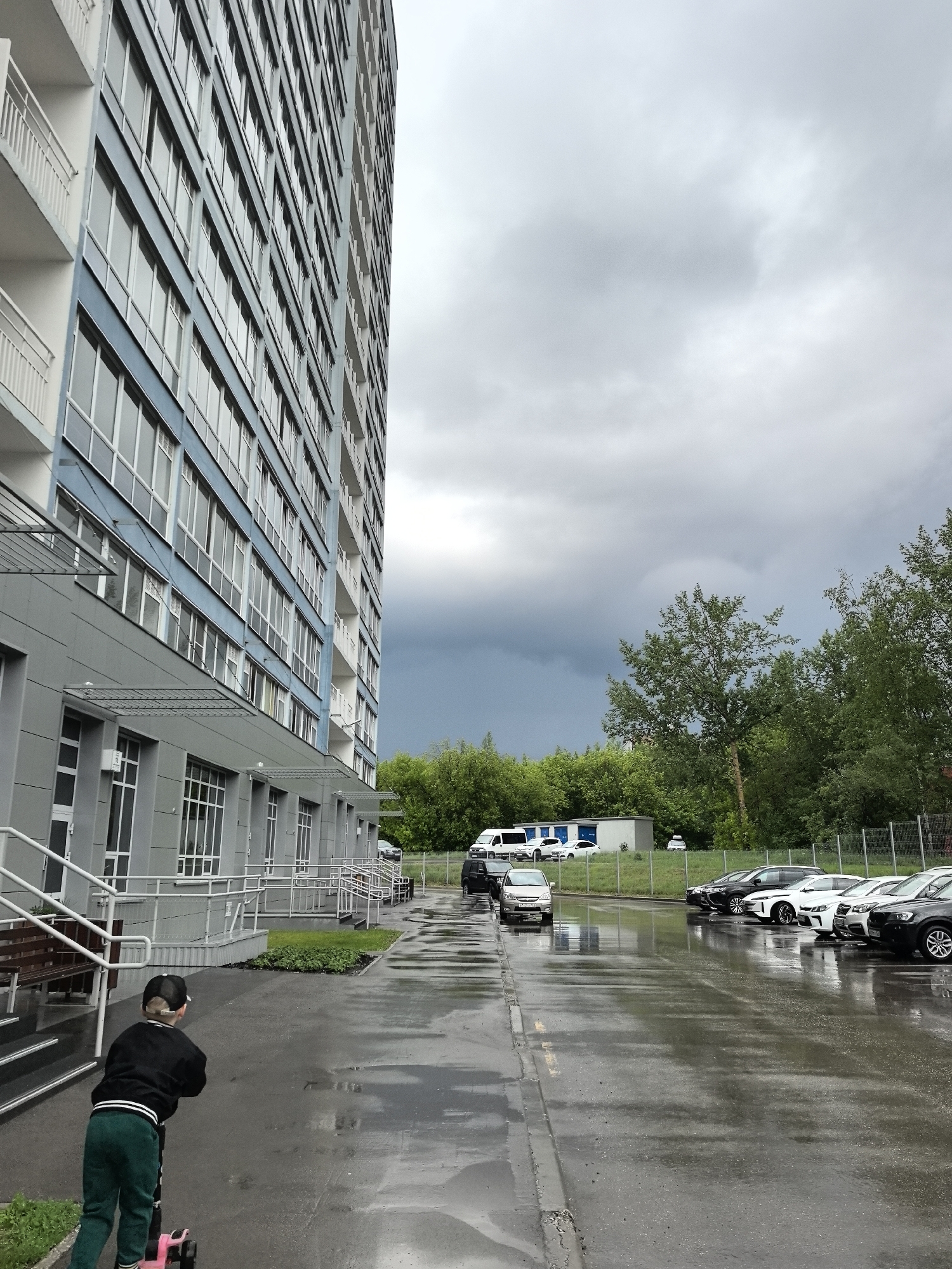 A low-angle, medium shot shows the exterior of a multi-story building on a wet day in Perm, Russia. A young child in a black jacket, green pants, and a baseball cap rides a scooter on the sidewalk in front of the building. The building has a gray facade with blue accents and numerous windows. A metal railing borders the sidewalk. Behind the building, trees line the horizon under a cloudy, dark sky. Multiple cars are parked in a lot behind a fence. A small white building is visible in the background. The ground is wet, reflecting the sky and surrounding objects.