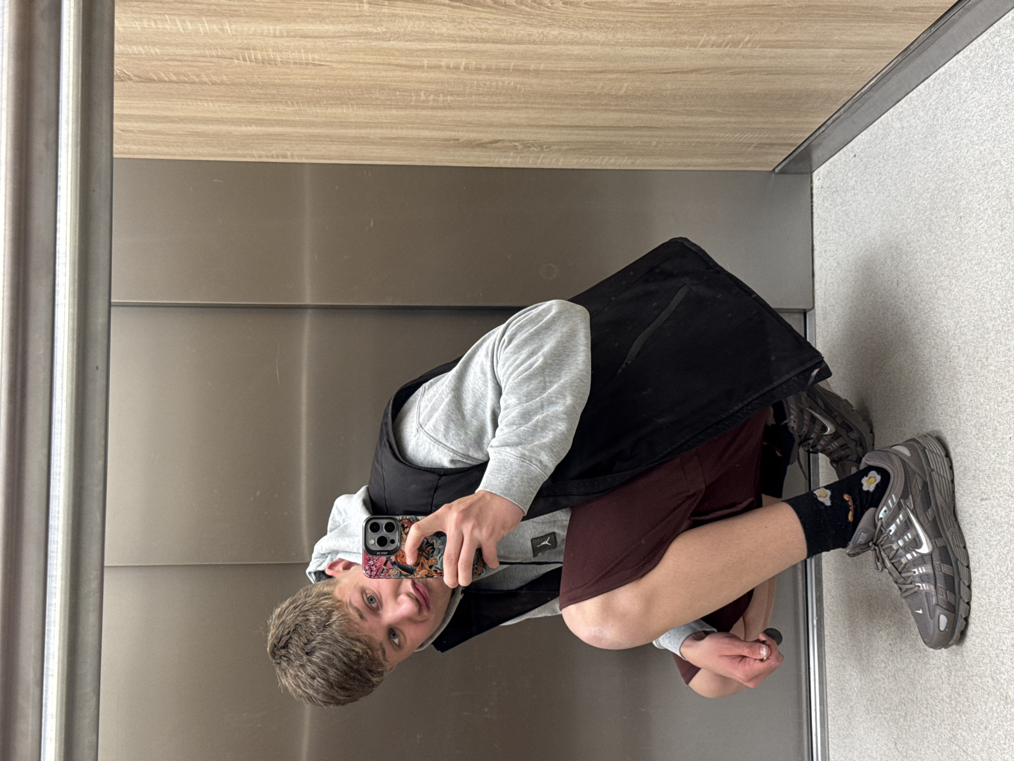 The provided image is rotated 90 degrees counter-clockwise and depicts a young man taking a mirror selfie indoors. The subject, a male in his late teens or early twenties, has light brown hair and blue eyes, looking directly at the camera with a neutral expression.
He is dressed in a grey hooded sweatshirt featuring a small dark logo (resembling the Jordan Jumpman), a black vest worn over the hoodie, and maroon shorts. His black socks display a playful pattern of white fried eggs and a bacon-like strip. He is wearing dark grey athletic shoes, identified as Nike by a visible silver swoosh. He holds a silver iPhone, likely a Pro model, encased in a colorful, intricately patterned phone case with faint text, possibly "BY COOP," near the camera.
The setting is an interior space, possibly an elevator or a modern room, consistent with a location in Plovdiv, Bulgaria. The background behind the subject is a reflective dark grey surface, functioning as the mirror for the selfie. Above this, the wall transitions to light-toned wood paneling. A vertical metallic trim is visible on the left side. The floor is a light grey, textured material, possibly carpet. The lighting appears to be bright and even, typical of indoor artificial illumination. The man's posture is a slight crouch, suggesting a casual selfie pose.