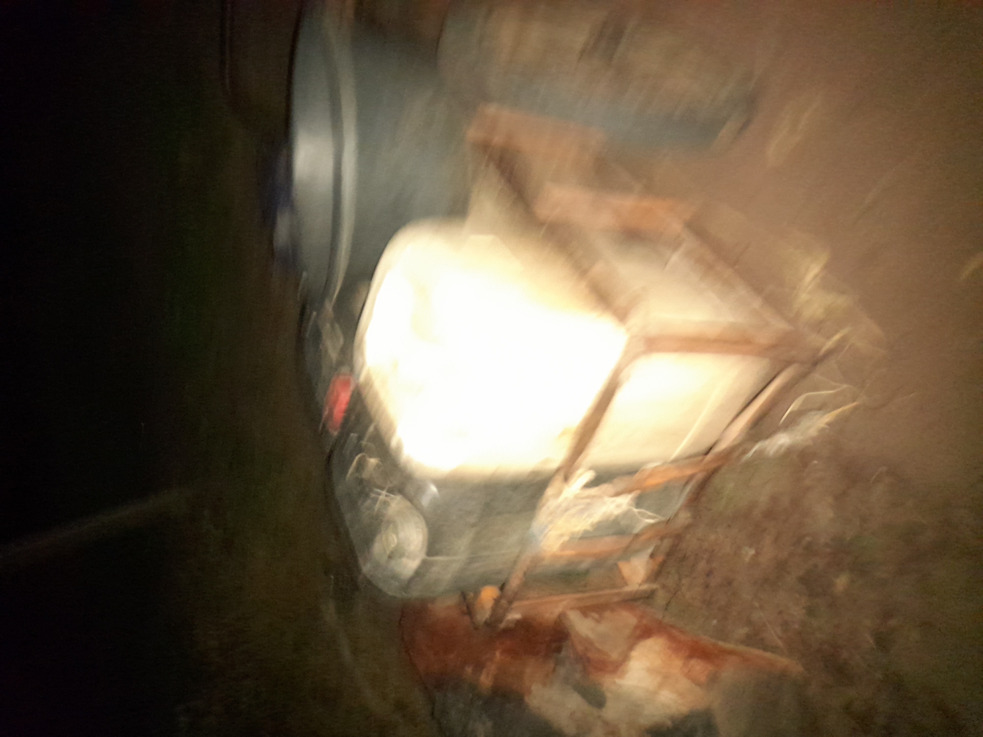 In Ibi, Nigeria, a blurry, night-time image captures a scene of what appears to be a makeshift transport setup. Two large, dark containers are centrally positioned. The one on the left is partially obscured and has a silver lid. To its right, a lighter-colored container, possibly plastic, is stacked on top and illuminated by a bright light source, casting a strong glare. These containers are resting on a wooden frame, which is itself situated on a rough, dirt-like surface. A dark, circular object, possibly a tire, is visible to the far right of the frame, and some reddish-brown material is present on the left side of the image, near the wooden frame. The background is dark and indistinct, suggesting an outdoor, dimly lit environment. The overall image quality is poor due to motion blur and low light conditions.