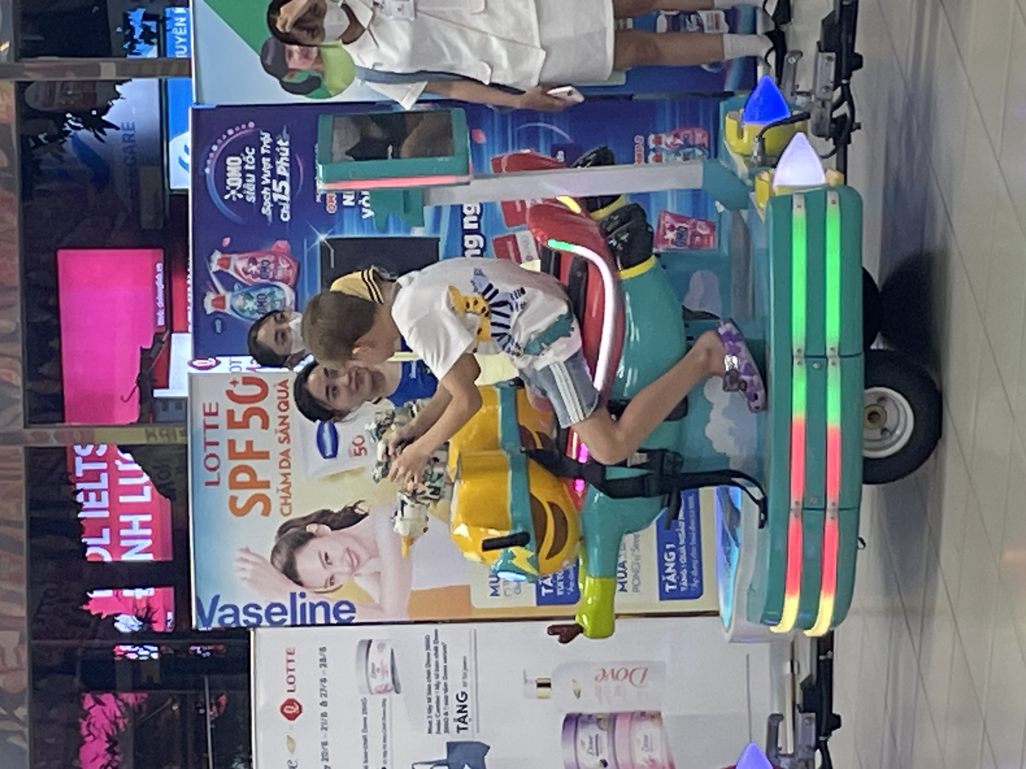 The image depicts an indoor scene, likely a commercial establishment or shopping mall in Thuận An, Vietnam, characterized by bright lighting and colorful advertisements. In the foreground, a child is seated on a multi-colored amusement ride that resembles a shooting game. The ride is green and yellow with red accents and features LED lighting around its base. The child is wearing a white t-shirt with a graphic print and shorts, and appears to be operating a toy gun.
Behind the child, several other individuals are visible. To the left, a young woman wearing a blue t-shirt is partially obscured. To the right, a woman in a white nurse-like uniform and a blue backpack stands, holding a mobile phone. Other individuals are seen in the background, some wearing face masks.
The environment includes large promotional banners and screens. One banner features a woman with the text "Vaseline SPF 50" and "LOTTE". Another banner displays images of laundry detergent bottles with Vietnamese text. The overall impression is of a vibrant, family-friendly entertainment or retail space. The floor is tiled with a light grey color.