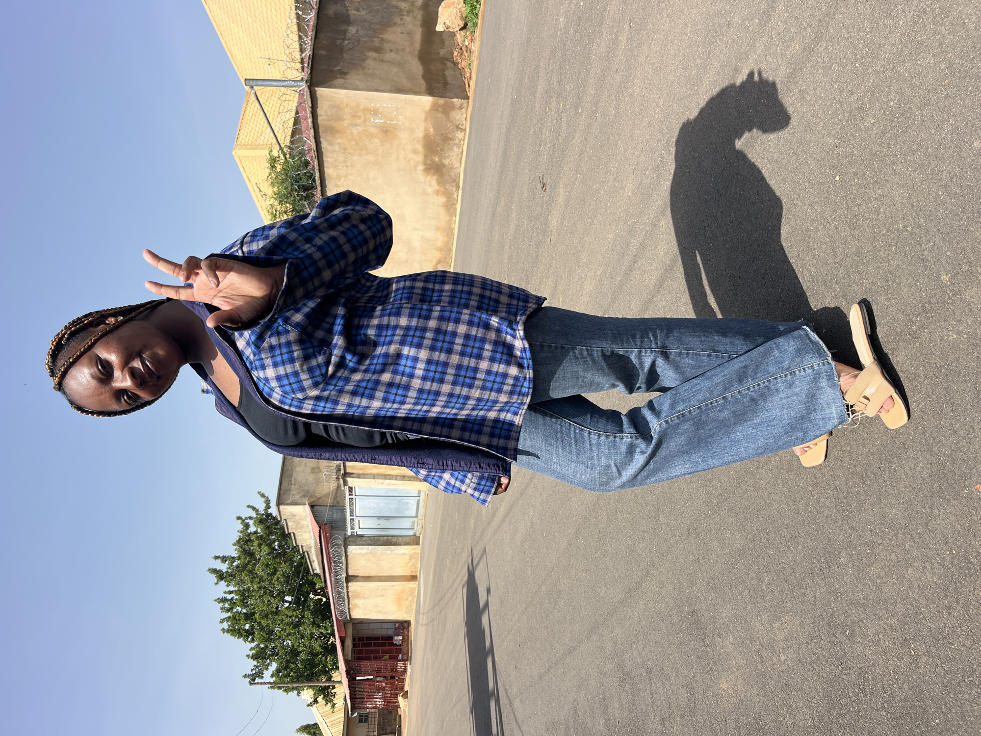 A full-body, outdoor, medium-shot photograph captures a woman standing on an asphalt road under a clear blue sky. She is positioned at an angle, facing the camera, with her left shoulder forward. Her hair is braided into dark brown cornrows. She is wearing a blue and gray plaid unbuttoned shirt over a black top, and loose-fitting blue jeans. Her left hand is raised, with her index and middle fingers extended in a peace sign. She is wearing beige slide sandals. The background features a row of low-rise buildings with tan walls and some with red metal gates or doors. A barbed wire fence runs along the top of some of the walls. Trees are visible behind the buildings. The sun casts a distinct shadow of the woman on the road to her right. The scene appears to be a residential street in an urban or semi-urban environment.