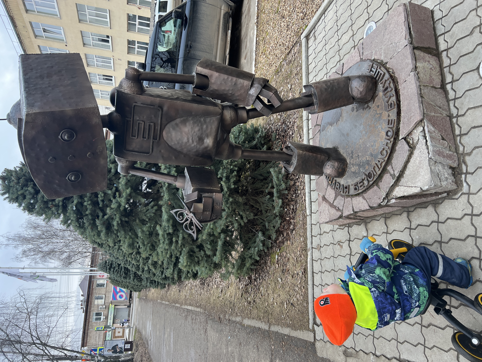 A statue of a robot made of dark metal stands on a stone base with a circular metal plate at the city of Chaikovsky, Russia. Inscribed text is visible on the plate. A child wearing a bright orange hat, blue and green patterned jacket, and blue pants rides a yellow and black tricycle next to the statue. A green evergreen tree is next to the statue, and a sidewalk runs parallel to the tree and statue. Buildings are visible in the background. A dark SUV is parked behind the statue.