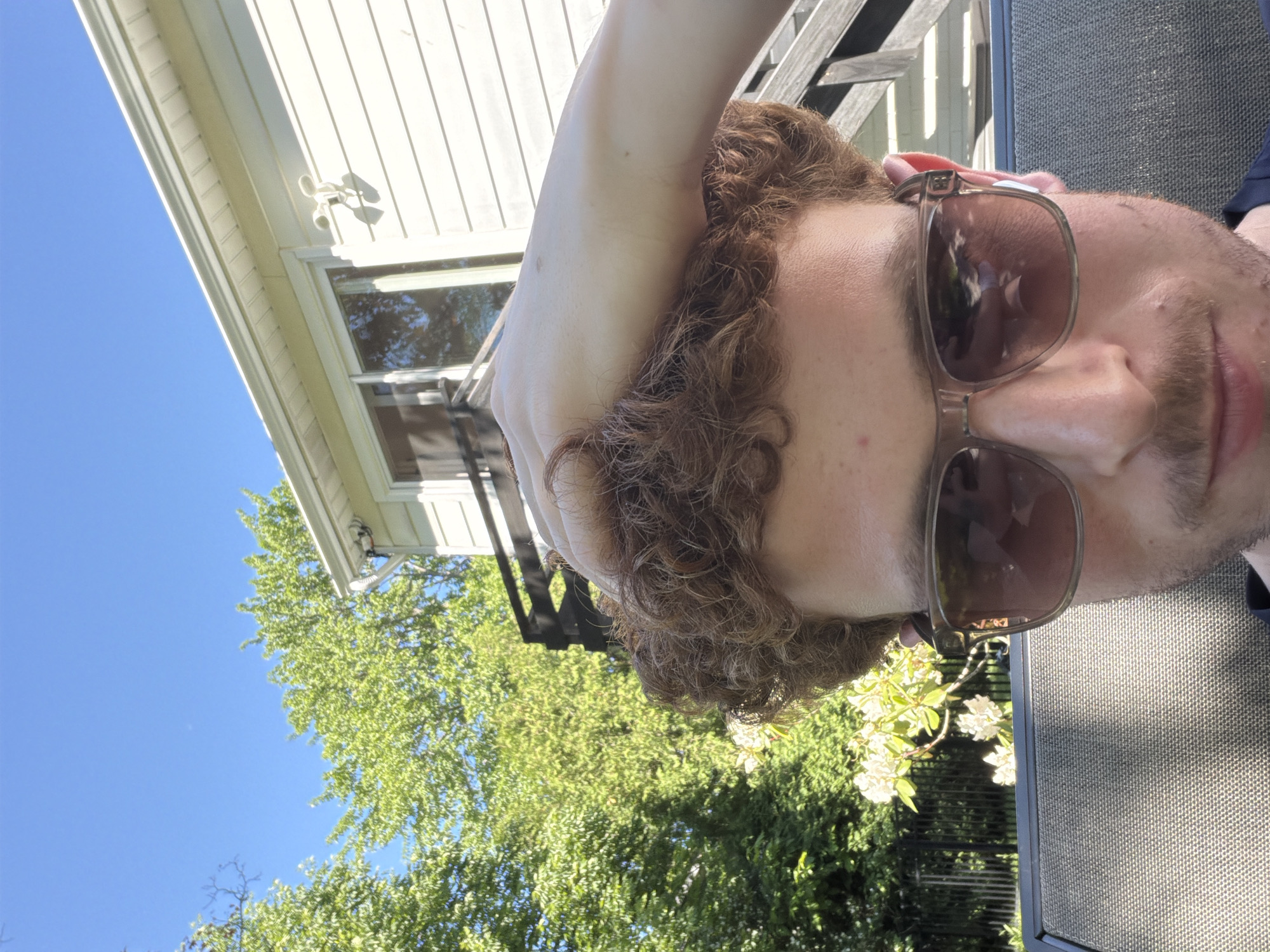 A close-up, angled outdoor shot features a man with curly brown hair and facial stubble, wearing dark-tinted sunglasses. His left arm is raised over his head, suggesting a relaxed posture. He appears to be lounging outdoors, possibly on a patio or deck.
In the background, a light-colored house with horizontal siding and a window is visible, along with a white outdoor fixture (possibly a light or security camera) mounted on its soffit. Lush green trees and foliage, accented by some white flowers, fill the midground, set against a bright, clear blue sky. Part of a dark, mesh-patterned outdoor furniture piece is visible to the right. The strong sunlight and clear sky indicate a sunny day, likely late morning or afternoon. The overall impression is one of leisure in a residential setting in Pepper Pike, United States of America. No visible text is present.