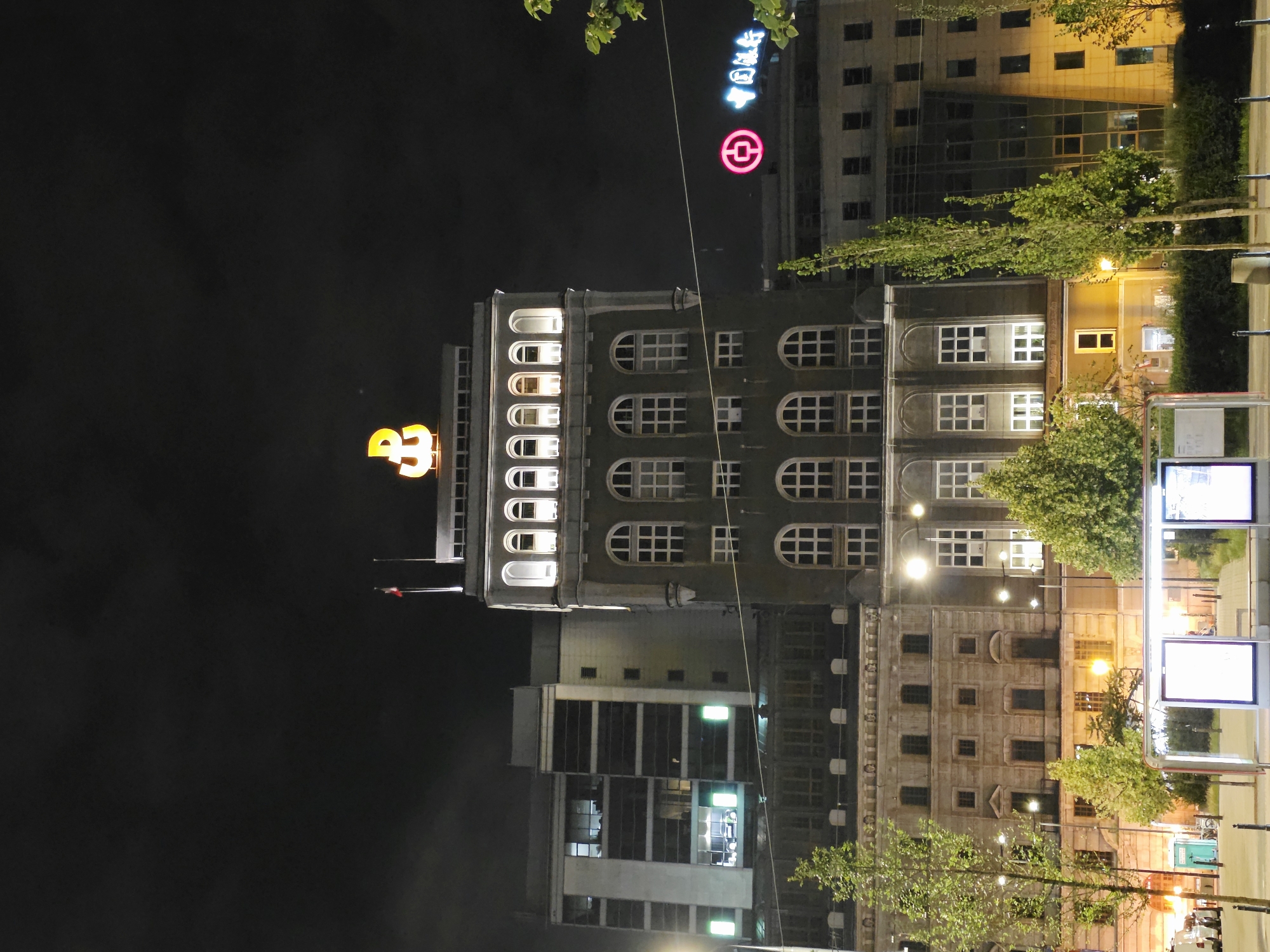 The image depicts a nighttime urban scene in Warsaw, Poland, focusing on a large, imposing building with illuminated windows and signage. The building features a distinctive yellow neon symbol resembling an anchor or a stylized 'P' at its peak, which is the emblem of the Polish State Railways (PKP) in Warsaw. The architecture is a blend of older, classical elements with modern additions. The lower floors show a more ornate, stone facade with arched windows, while the upper sections and adjacent buildings appear more contemporary with large glass panels and uniform windows.
Several trees, some illuminated by streetlights, line the foreground and midground, suggesting a public space or plaza. A modern bus stop shelter with illuminated advertising panels is visible on the left, indicating public transportation infrastructure. The sky is dark and cloudy, typical of an evening in the city. There are no visible people clearly depicted in the scene, though the illuminated windows suggest occupancy within the buildings. The overall impression is one of a busy urban center at night, with prominent architectural features and commercial signage. To the right, a neon sign with Chinese characters and a circular logo is visible on another building, indicating a Chinese bank.