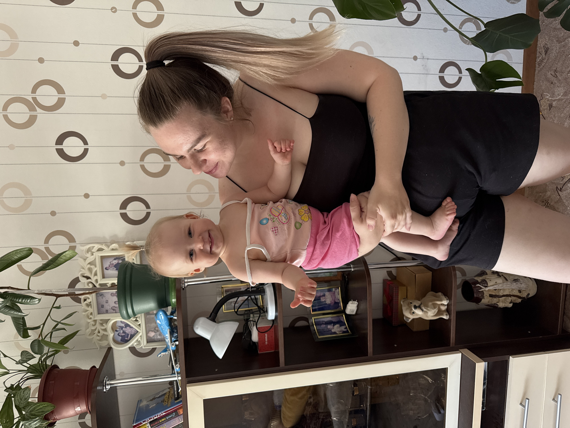 A fair-skinned woman with her blonde hair in a ponytail is holding a baby. She is wearing a black top and black shorts. The baby is wearing a pink outfit with a white trim. The woman is looking down at the baby with a smile. The background shows a wall with circular designs, a bookshelf with various objects, and two potted plants. The location is Chaikovsky, Russia.