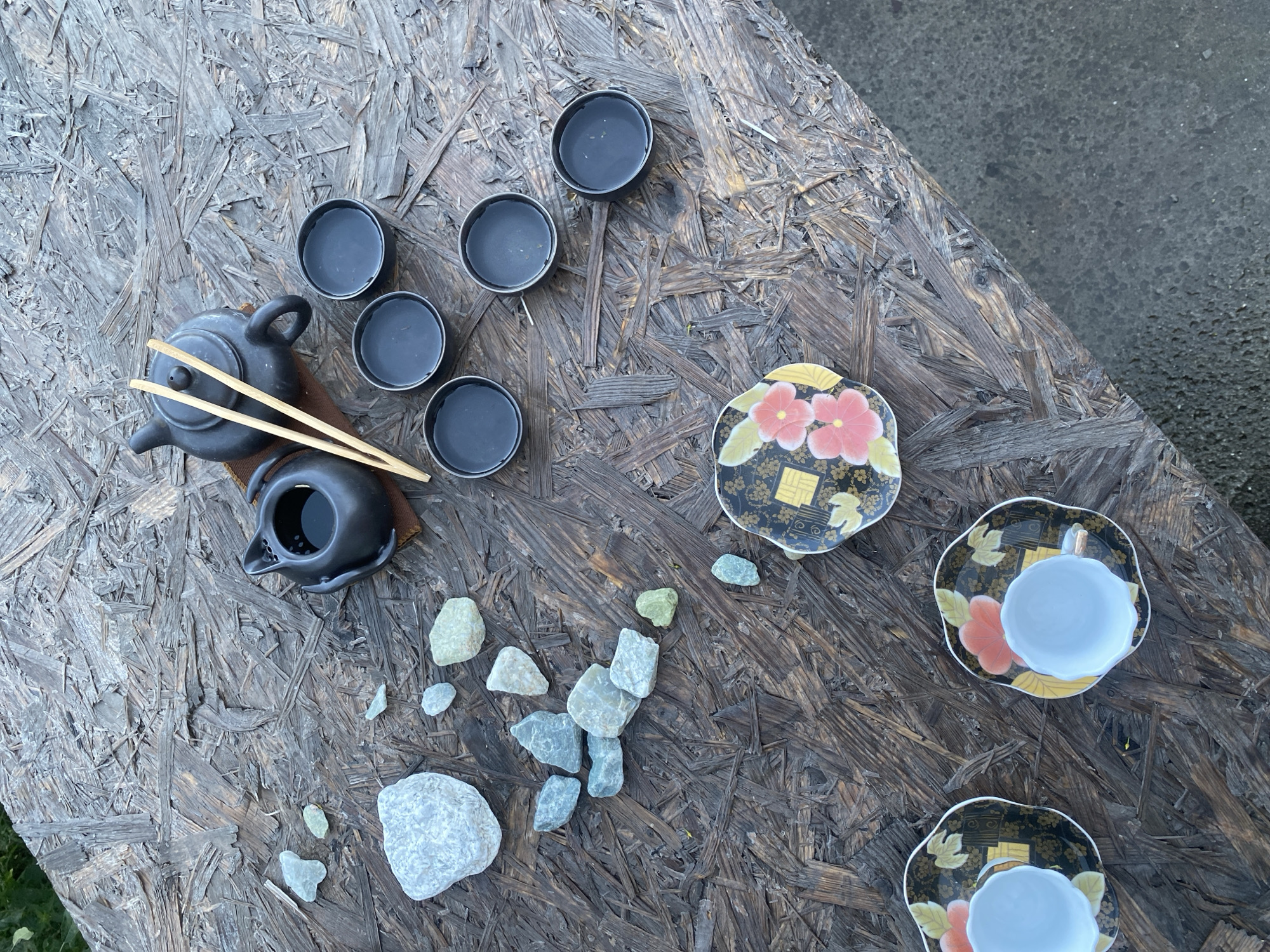 A top-down, medium shot captures a still life arrangement of a tea set and rocks on a weathered, textured wooden surface, possibly an outdoor table. The scene is set outdoors, with a hint of green foliage visible at the top edge, suggesting a natural or garden setting. The wooden surface appears to be made of compressed wood chips, with a rough and uneven texture.
In the upper right quadrant of the image, a dark gray teapot with a lid and a smaller dark gray pitcher are positioned on a small brown cloth. Two light-colored wooden chopsticks rest on the lid of the teapot. To the right of the teapot, a row of six dark, shallow cups are arranged, each filled with a dark liquid, presumably tea.
In the lower right quadrant, two small saucers with a black background and a floral pattern of pink and yellow flowers and leaves are visible. Each saucer holds a white teacup.
Scattered across the left and middle portions of the wooden surface are various rocks. The largest is a pale gray, smooth, rounded rock. To its right and below, a cluster of smaller, irregularly shaped rocks in shades of light green and gray are arranged.
The ground surface visible along the bottom right edge of the image is a dark, textured asphalt or concrete. There are no people visible in the image. The lighting suggests it is daytime, with soft shadows. The image does not provide any specific details that would indicate a location in Almaty, Kazakhstan, beyond it being an outdoor scene.